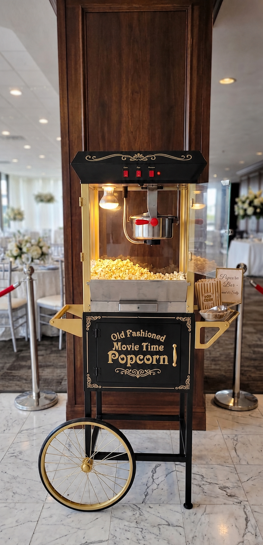 A vintage-style popcorn machine on a black cart with gold wheels set up in the luxury banquet ballroom at JPS Events at Regions Tower in Downtown Indianapolis, surrounded by reception tables with white floral arrangements.