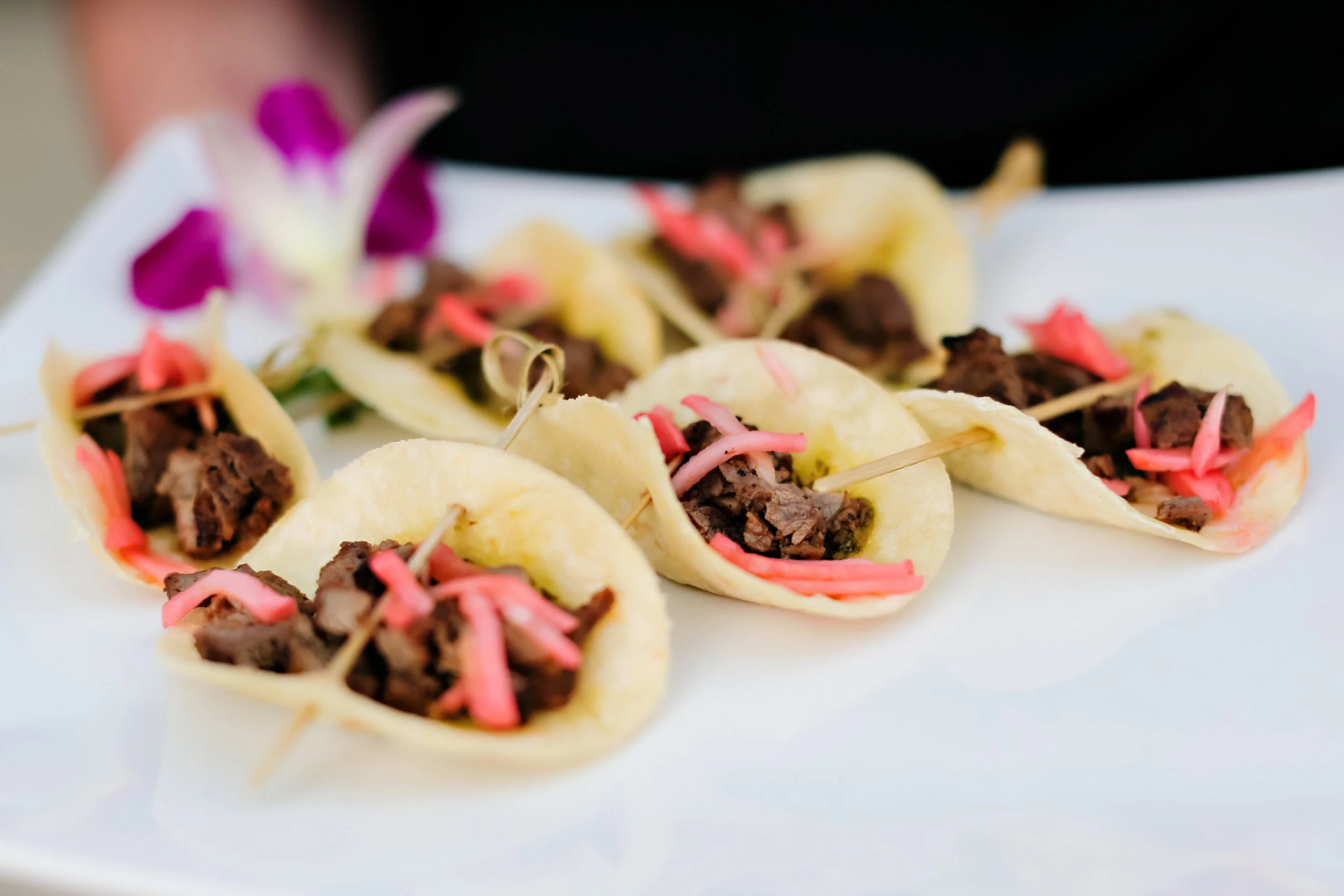 Mini tacos filled with cooked meat, topped with pink shredded garnish, served on a white plate.