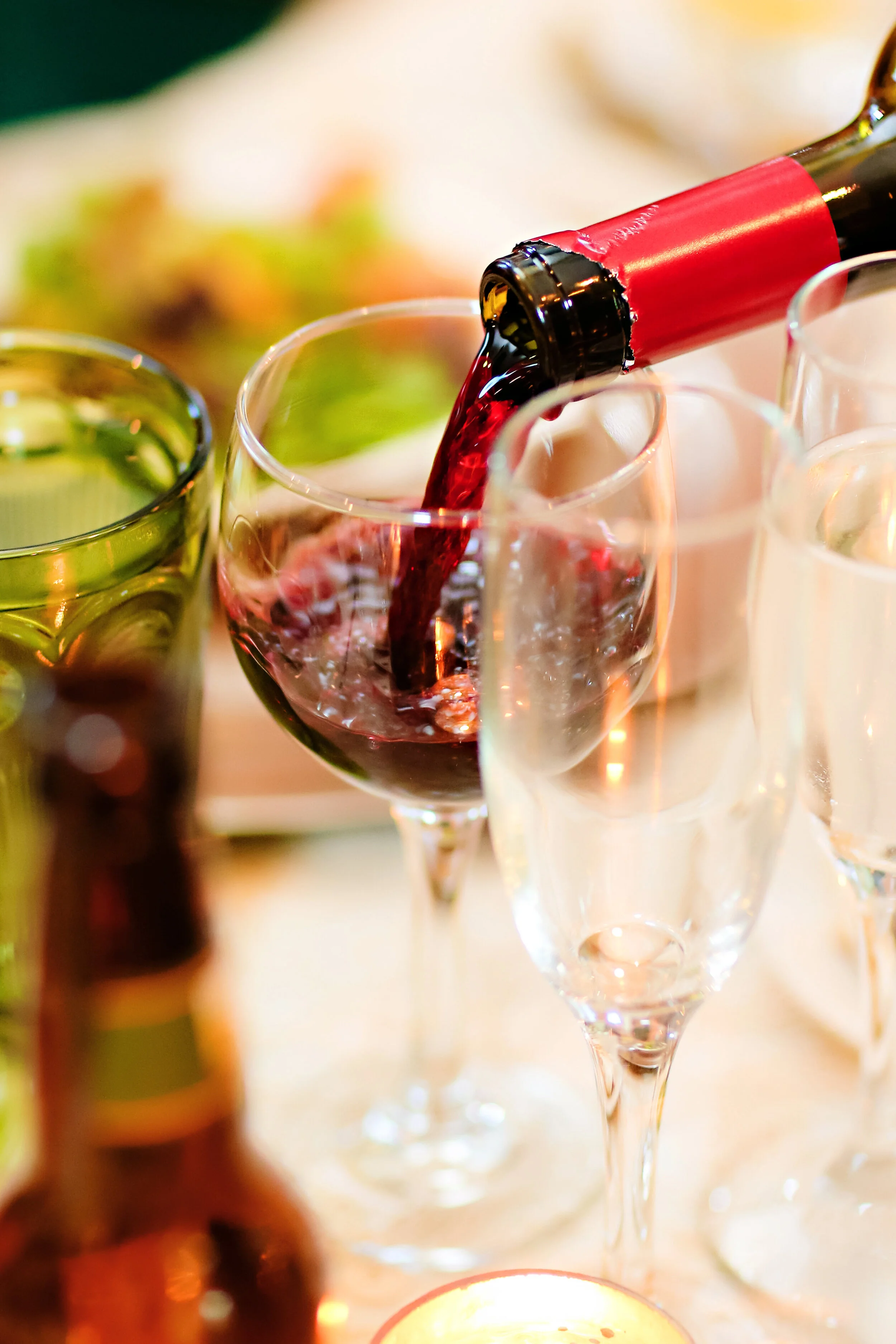 A glass of red wine being poured from a bottle with a red label, accompanied by other empty glasses and a candle