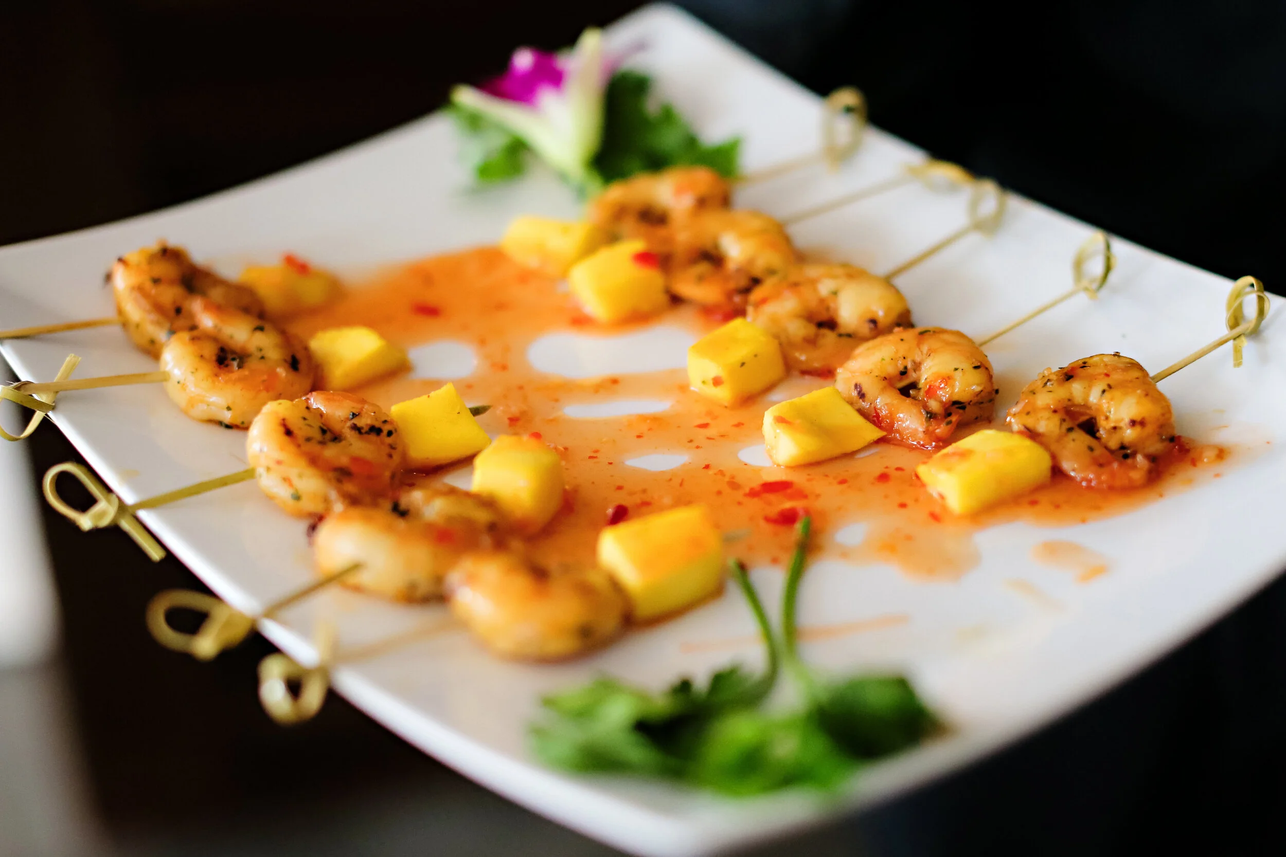Shrimp skewers on a white plate with pineapple chunks and a green garnish, with a dark background.