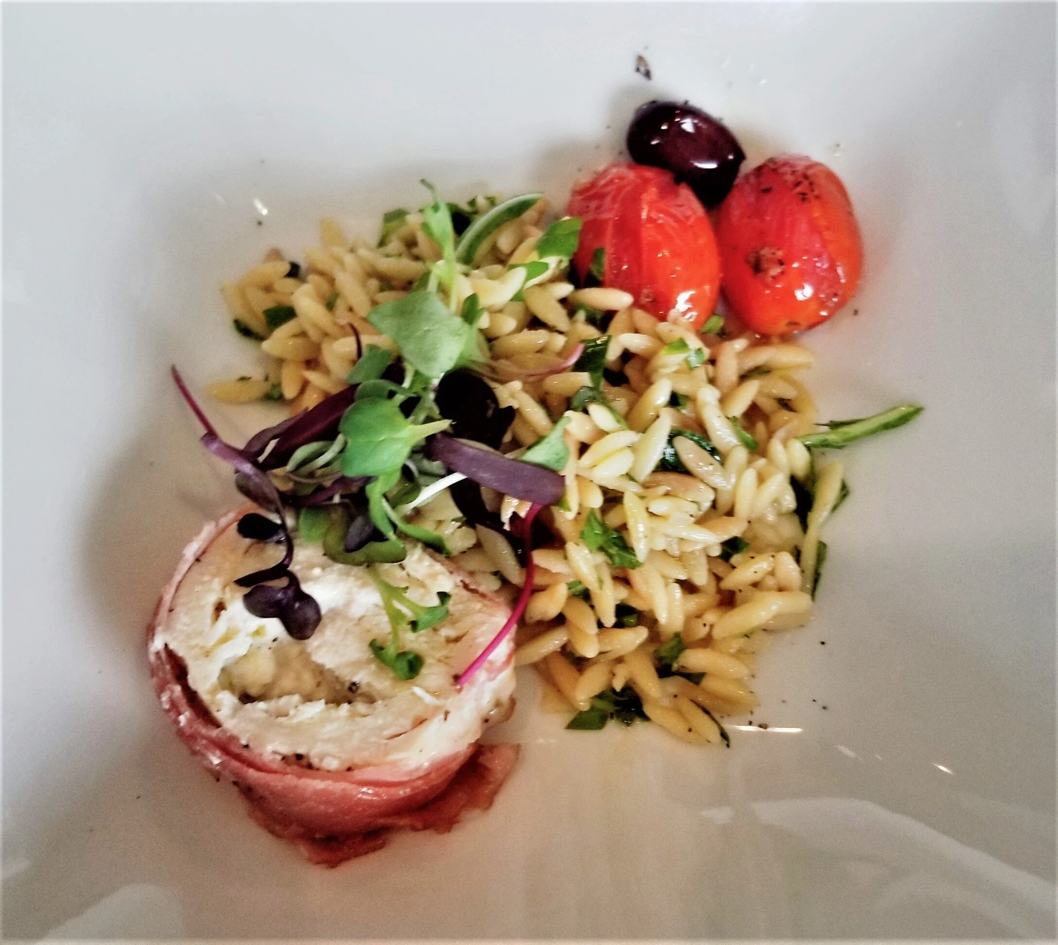 A bowl containing a stuffed lobster tail topped with creamy filling, served with orzo pasta salad garnished with fresh herbs and cherry tomatoes.