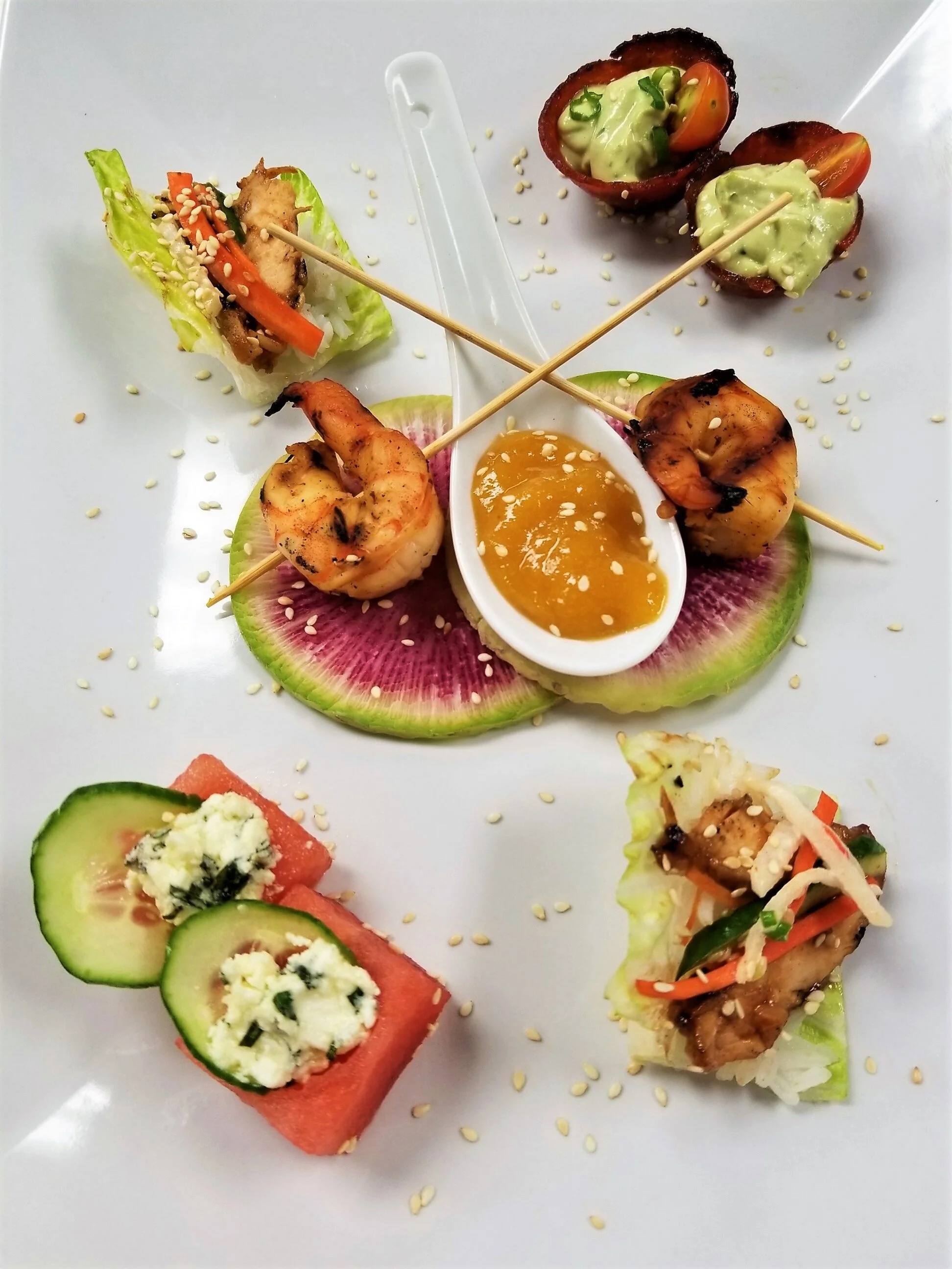 Assorted bite-sized sushi rolls and mini tacos on a white plate, garnished with sesame seeds.