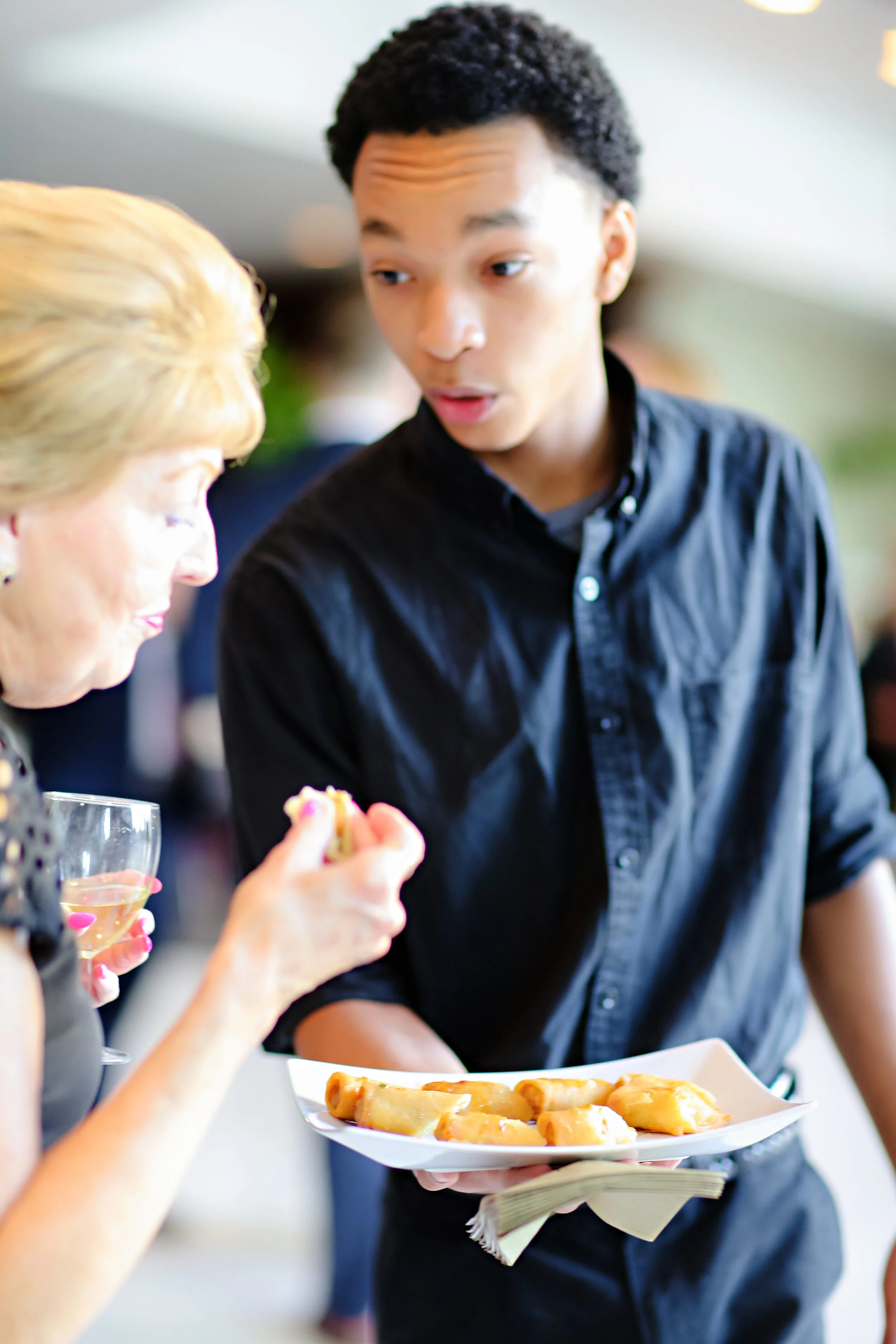 A guest being served delicious food by our luxury in-house catering at JPS Events at Regions Tower in Downtown Indianapolis, with a woman in the foreground holding a glass of wine and a plate of food at a wedding reception