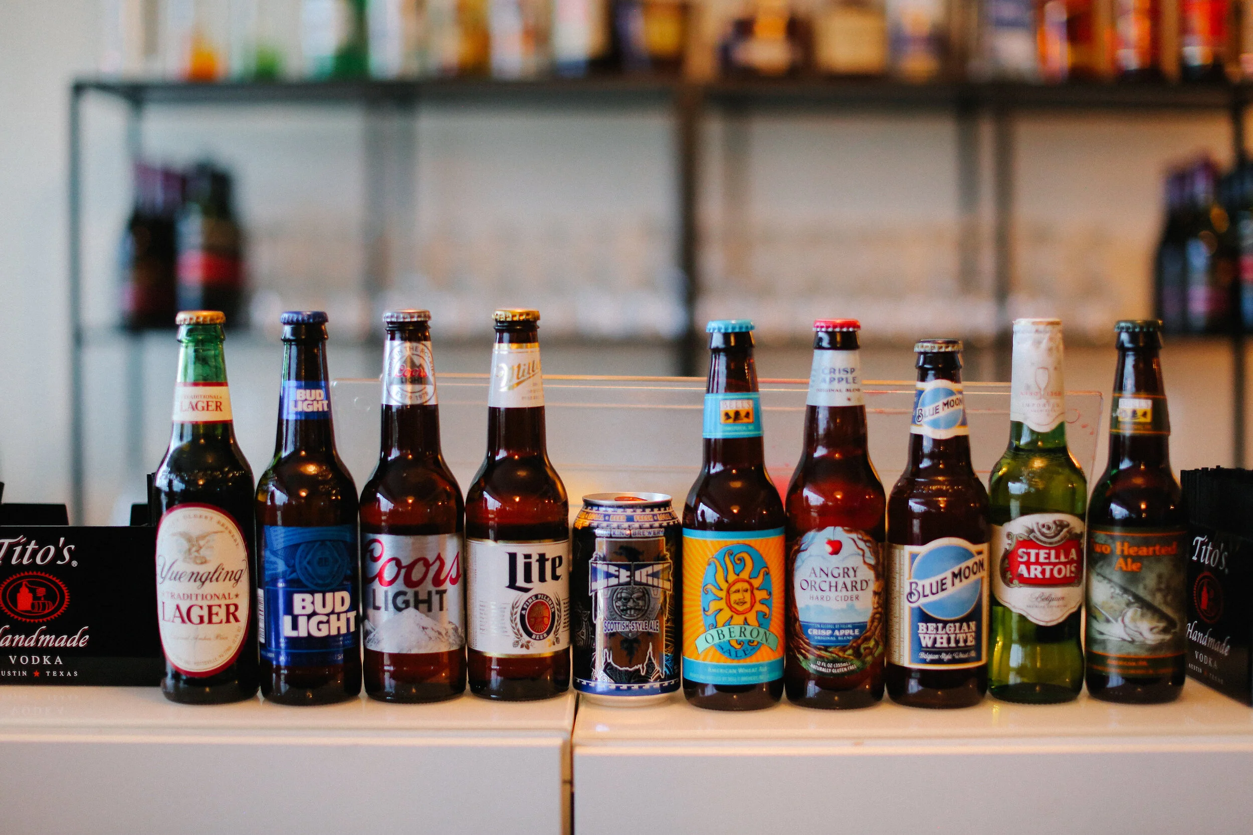 A display of various beer bottles on a bar counter with a blurred bookshelf in the background.