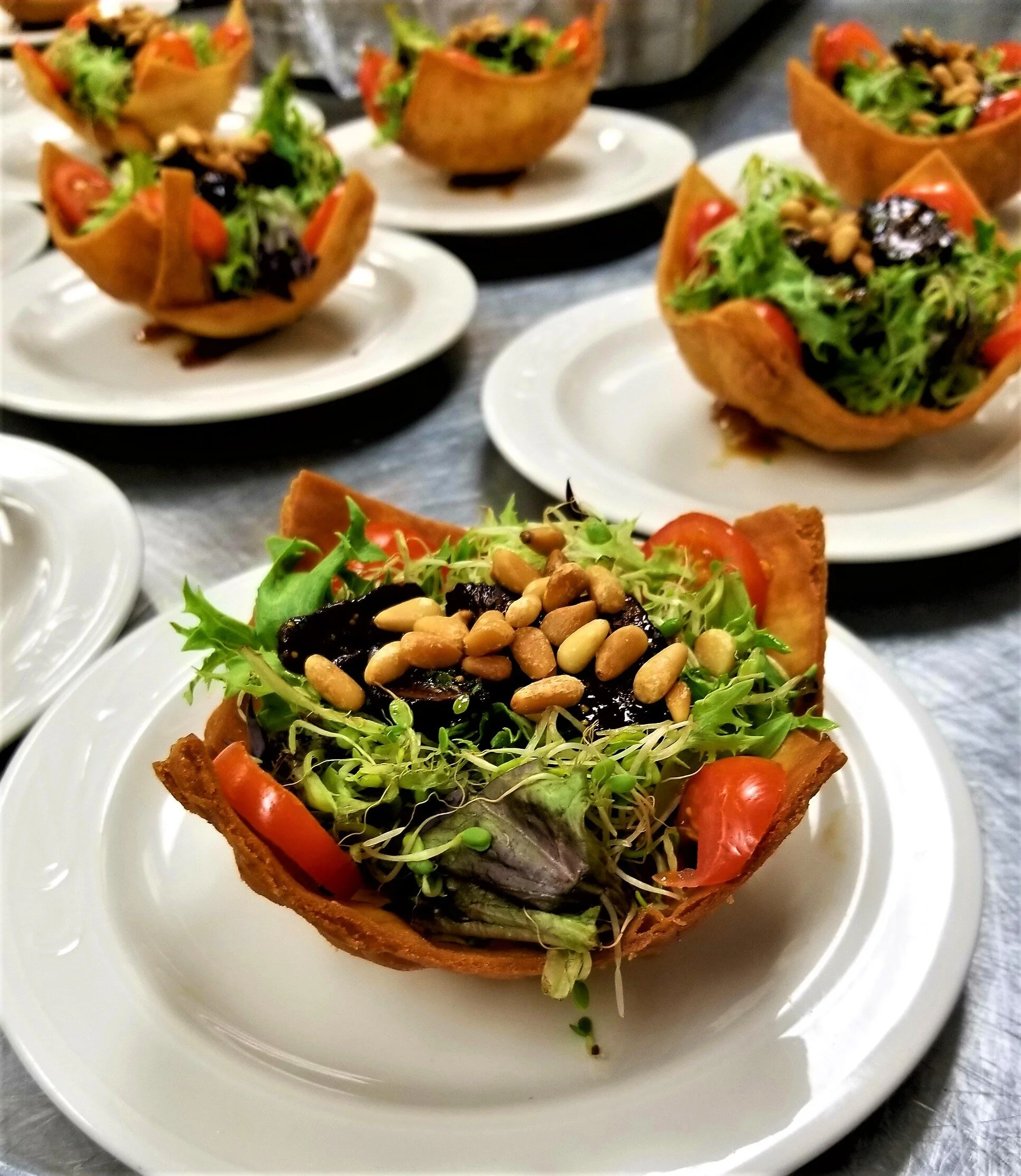 Bowl shaped crispy fried tortilla filled with lettuce, cherry tomatoes, black beans, pine nuts, and microgreens on a white plate.