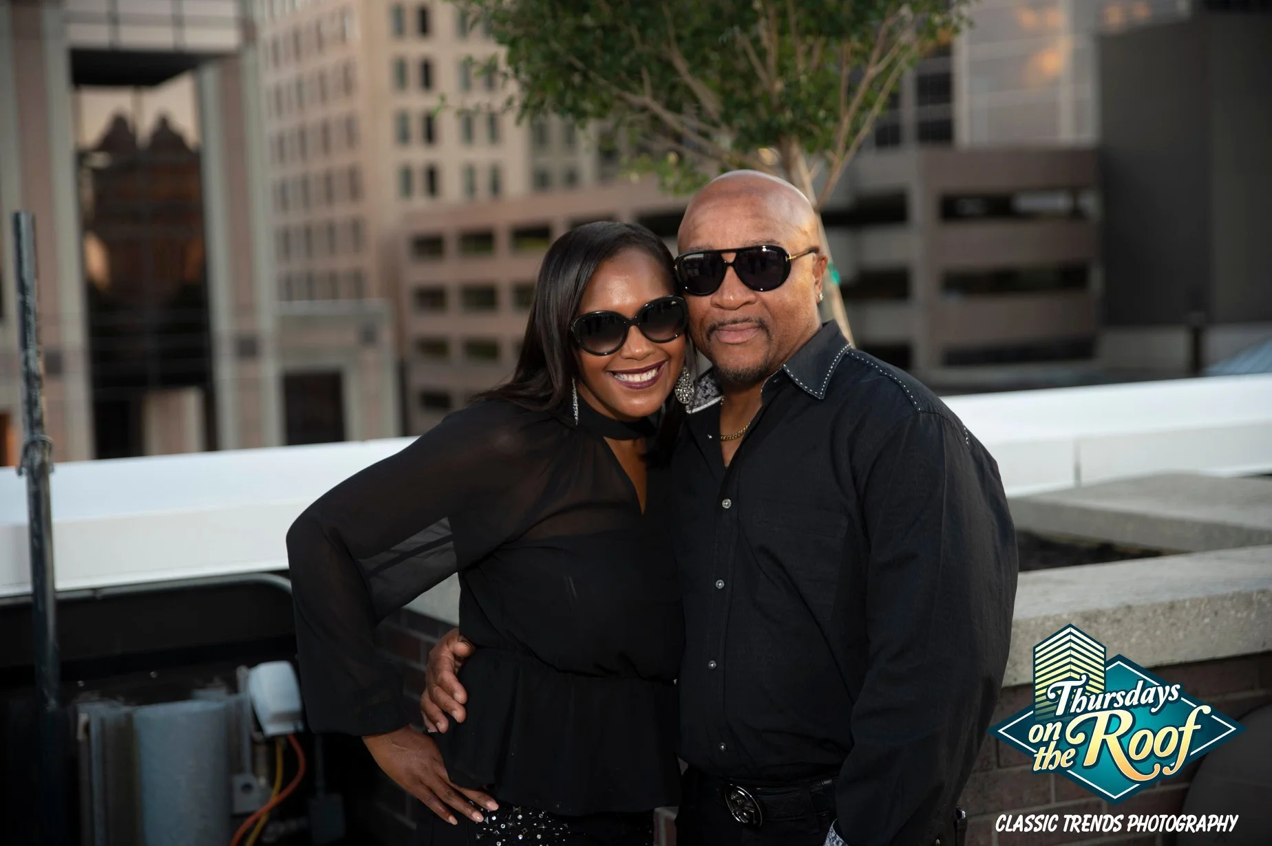 A smiling woman and man with sunglasses posing together during Thursdays on the Roof on the 5th floor terrace at JPS Events at Regions Tower in Downtown Indy, with an urban setting and skyline tall buildings in the background.  