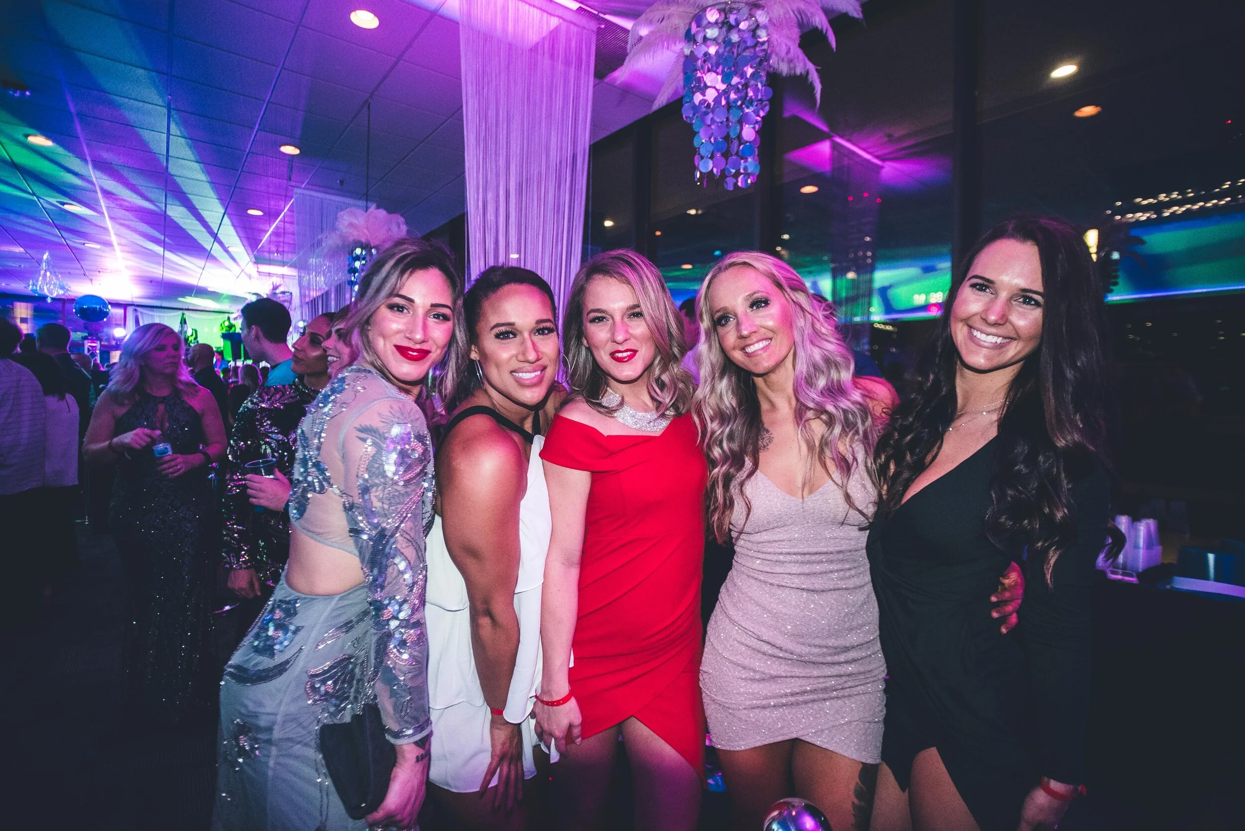 Five women dressed in elegant attire at a party or nightclub, with colorful lighting and decorations in the background at HOT NYE (formerly CÎROC the New Year) at JPS Events at Regions Tower in Downtown Indy.