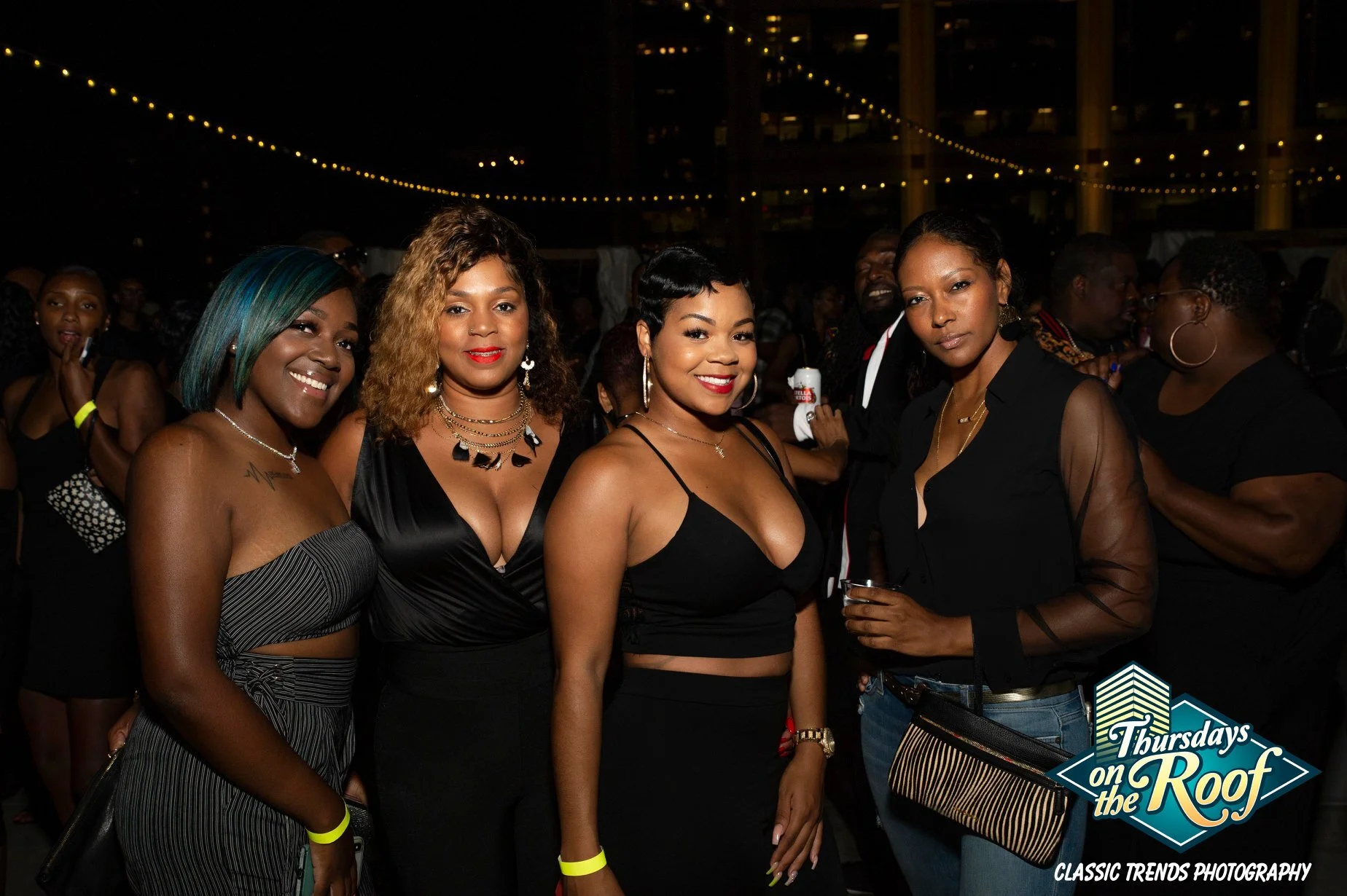 Group of women at Thursdays on the Roof, an rooftop nightclub event at JPS Events at Regions Tower in Downtown Indy, dressed in black, with string lights overhead and a logo for 'Thursdays on the Roof' in the lower right corner.  