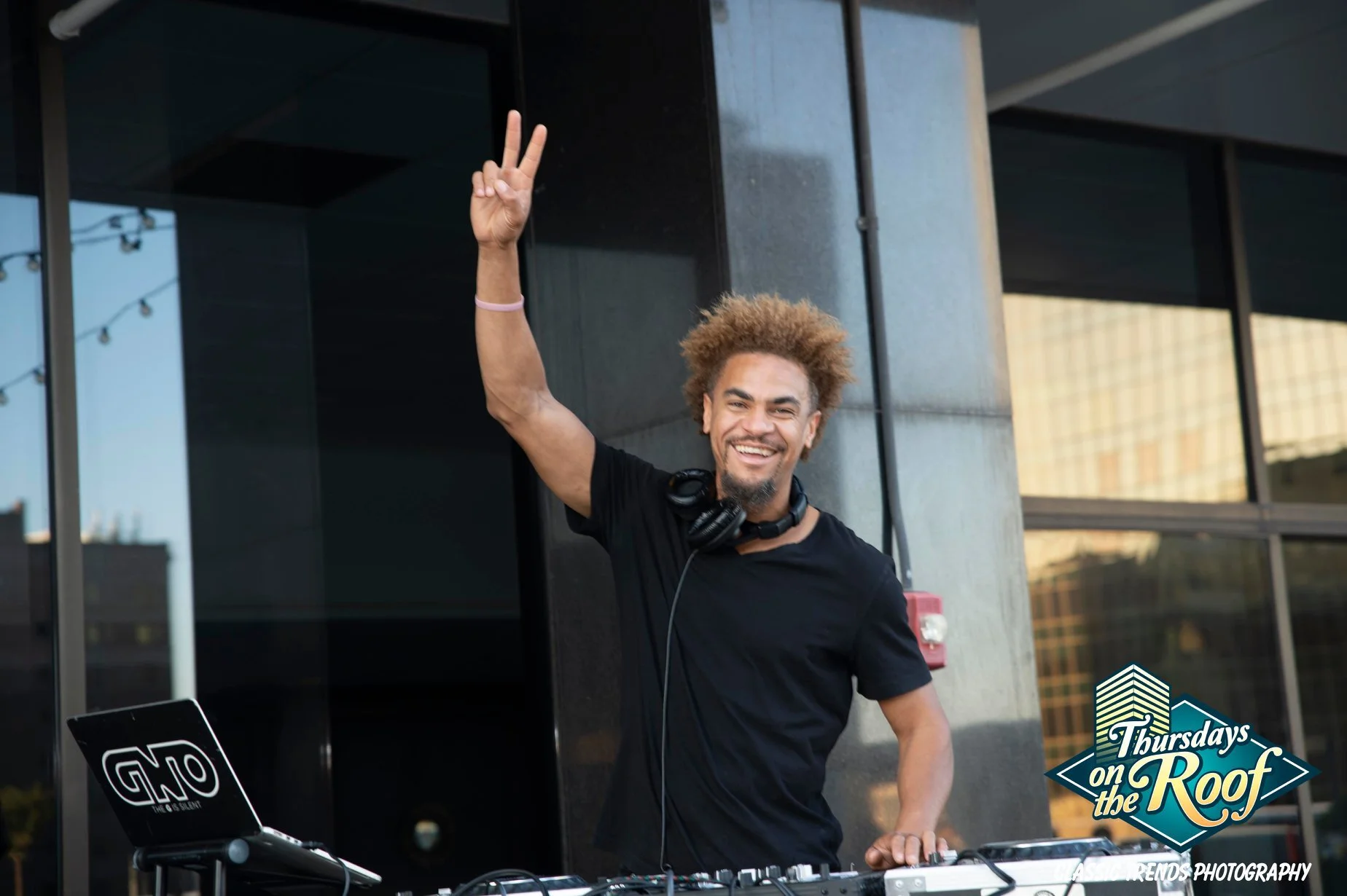 A smiling DJ wearing headphones and a black T-shirt raising his hand during Thursdays on the Roof at JPS Events at Regions Tower in Downtown Indy, an iconic nightlife mainstay with live DJs, food, and rooftop bars.  