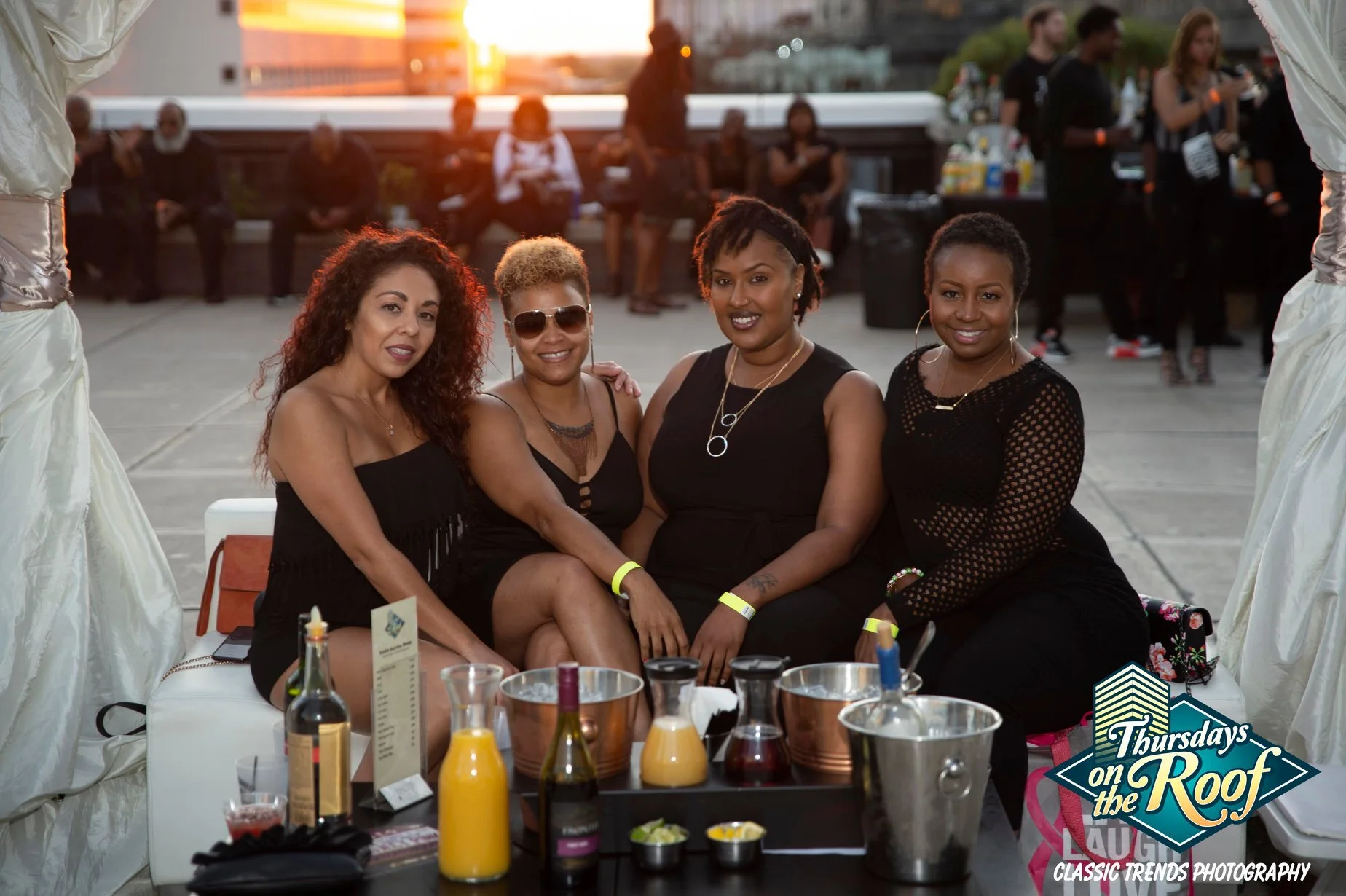 Four women sitting together at a private cabana on the 5th floor rooftop during sunset at Thursdays on the Roof at JPS Events in Downtown Indy, an iconic nightlife mainstay with live DJs, bottle service, food, and bars with city views.  