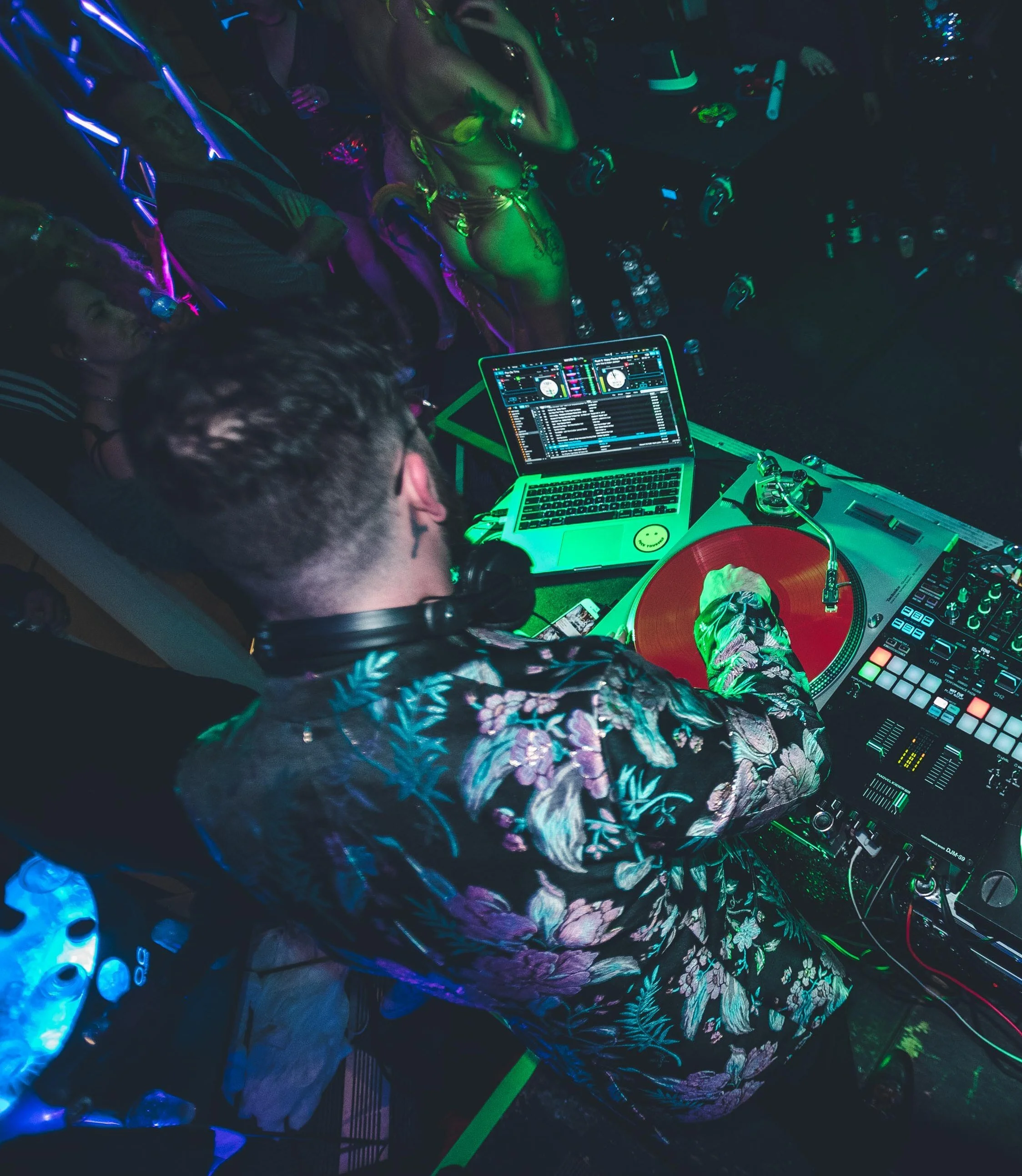A DJ mixes music at a party with people dancing around at HOT NYE (formerly CÎROC the New Year) at JPS Events at Regions Tower in Downtown Indy. The scene is dark with colorful neon lights, with drinks and bottles nearby.  