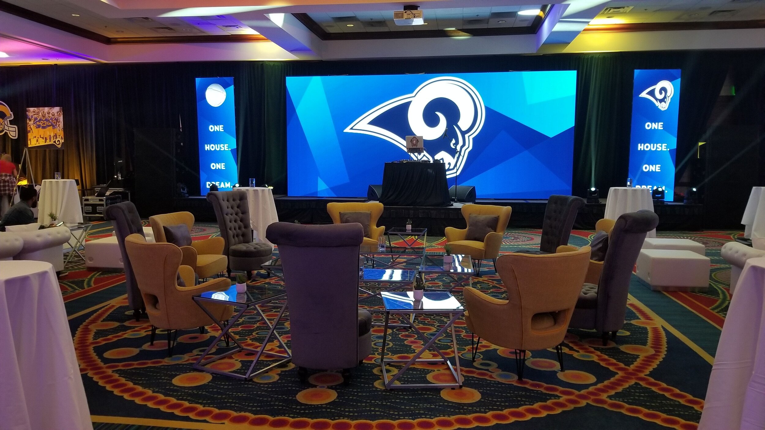 Produced by JPS Events: a spacious room with a stage decorated with LA Rams logos for the LA Rams Super Bowl after party. Vertical screens display the Rams logo and 'One House. One Dream.' with event seating and lighting accents.  
