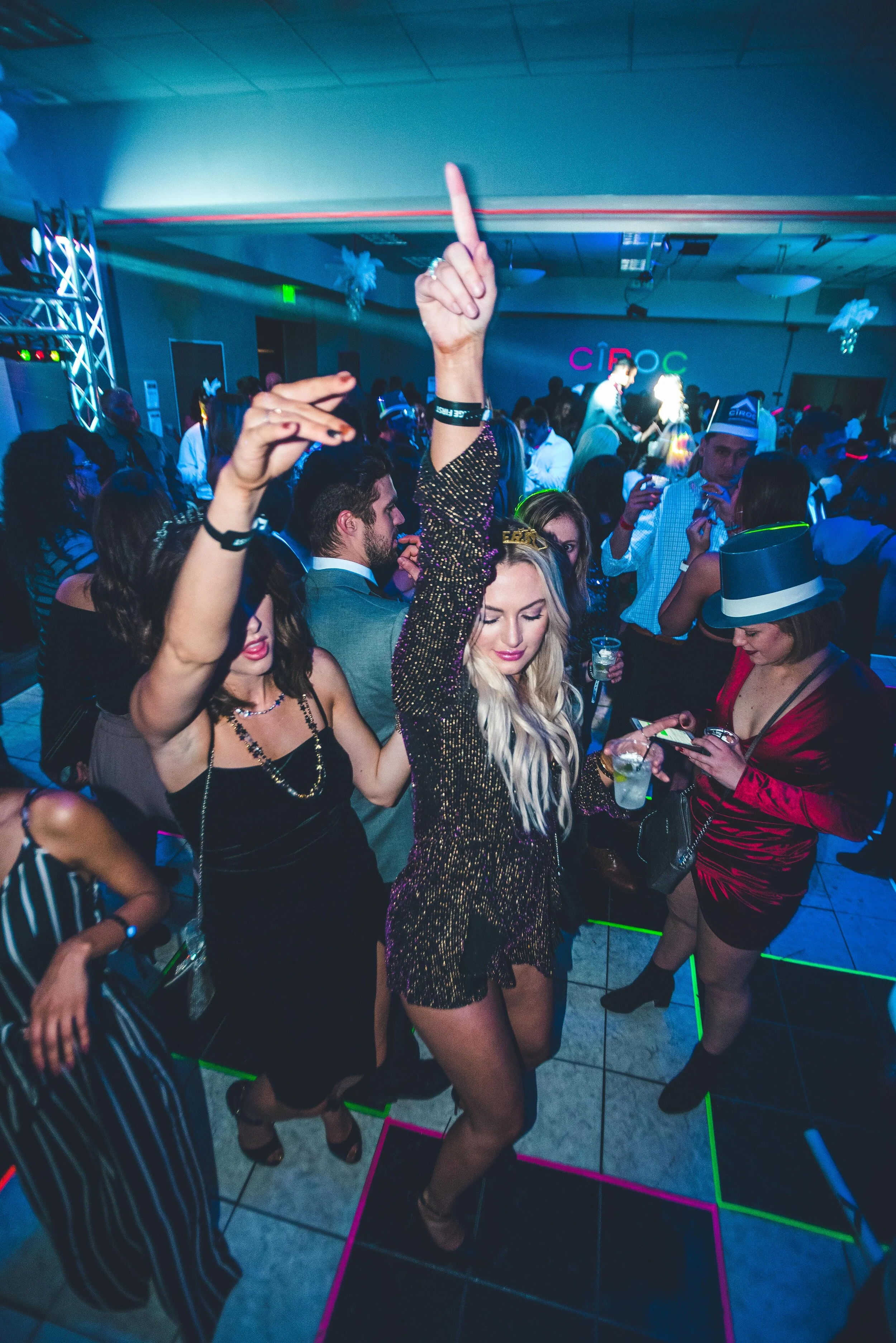 People dancing and socializing at HOT NYE (formerly CÎROC the New Year), a premier party at JPS Events at Regions Tower in Downtown Indianapolis, featuring vibrant nightclub lights, neon signs, and 360-degree skyline views.  