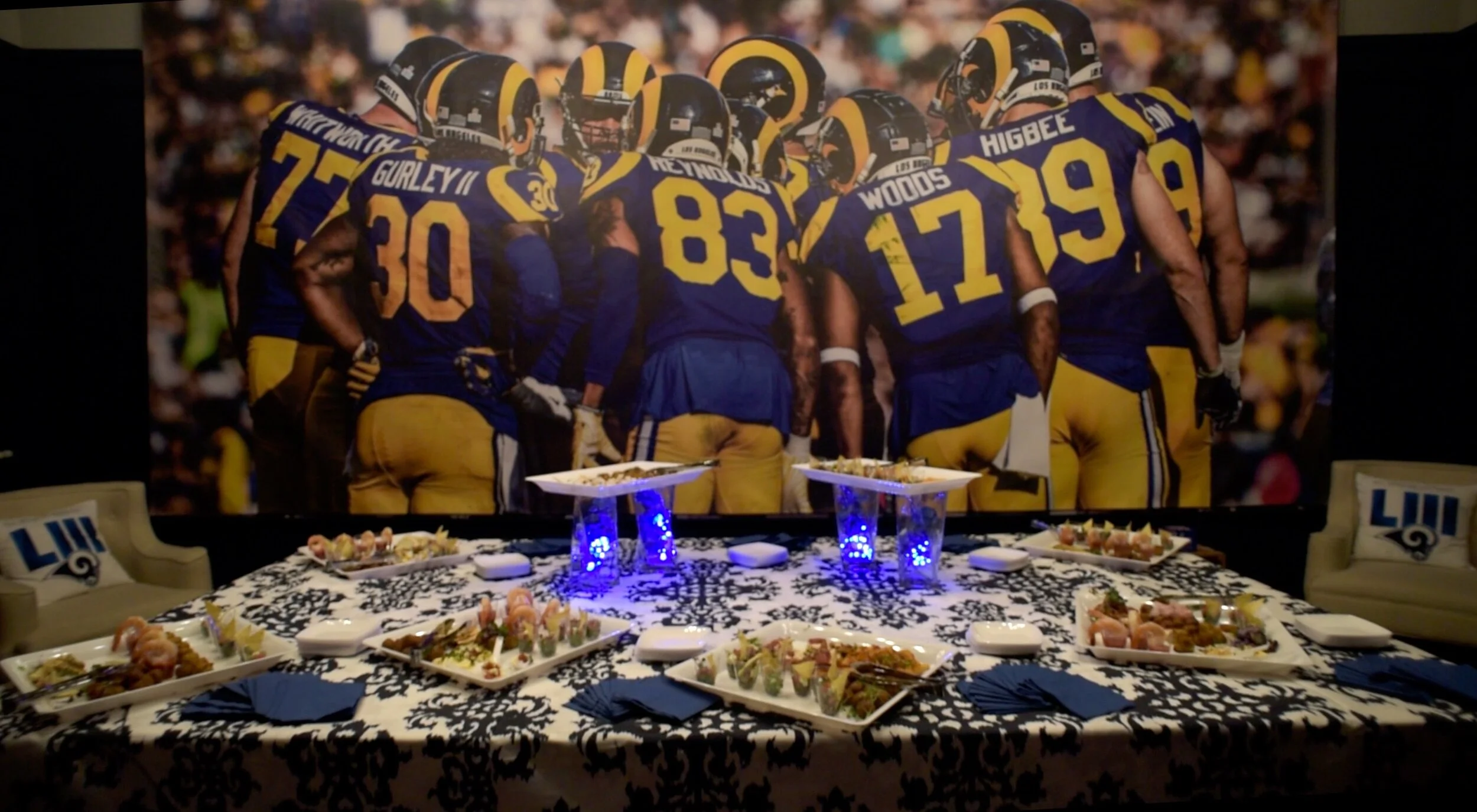 A table set with various appetizers and snacks in front of a large photo of Los Angeles Rams football players during a game, catered by luxury in-house catering for the LA Rams Super Bowl after party produced by JPS Events.  