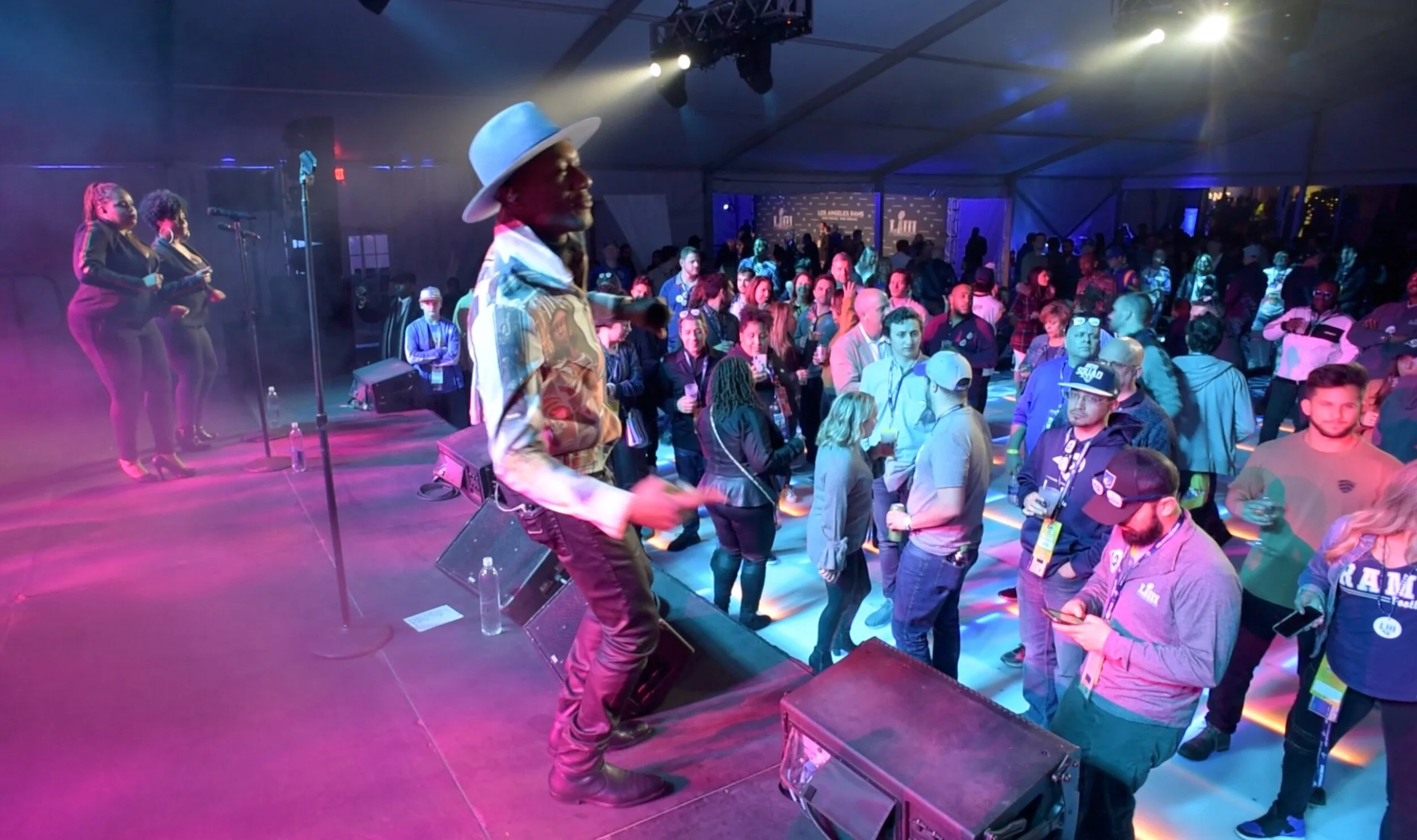 A live performance at a crowded event with a singer in a large hat singing on stage, and dancers in black outfits, produced by JPS Events for the LA Rams Super Bowl after party. Audience standing and dancing under colorful lights.  