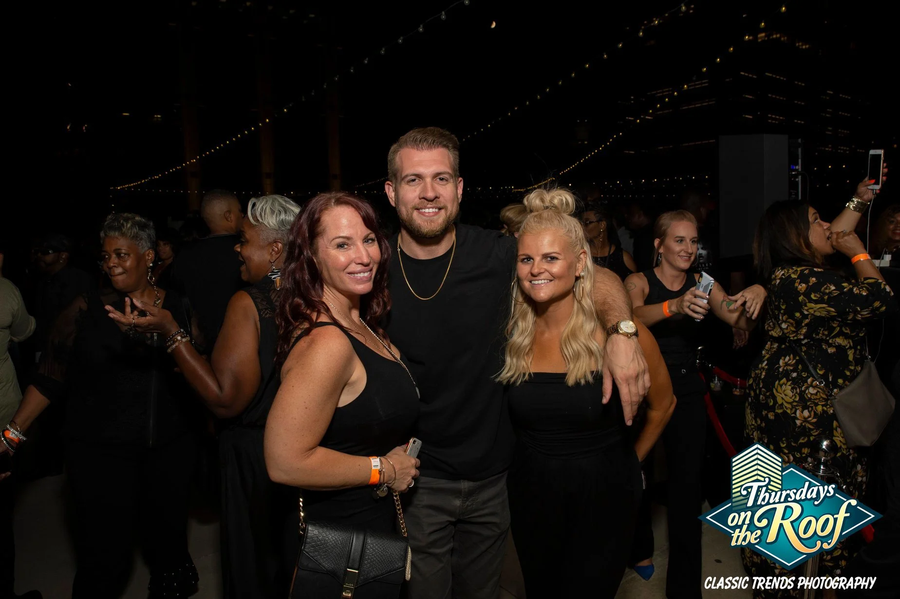 A group of four smiling people, two women and a man, standing together at a nighttime outdoor event with string lights overhead. at Thursdays on the Roof at JPS Events in Downtown Indy, an iconic nightlife mainstay with live DJs, bottle service, food