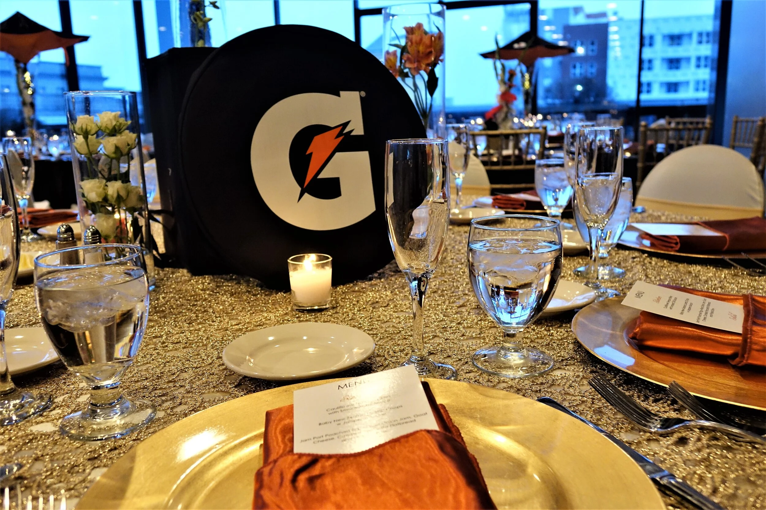 A banquet table set for an event with gold tablecloths, glassware, flowers, and a black Gatorade cooler. The table is decorated with a lit candle and place settings, with city buildings visible through large windows in the background.