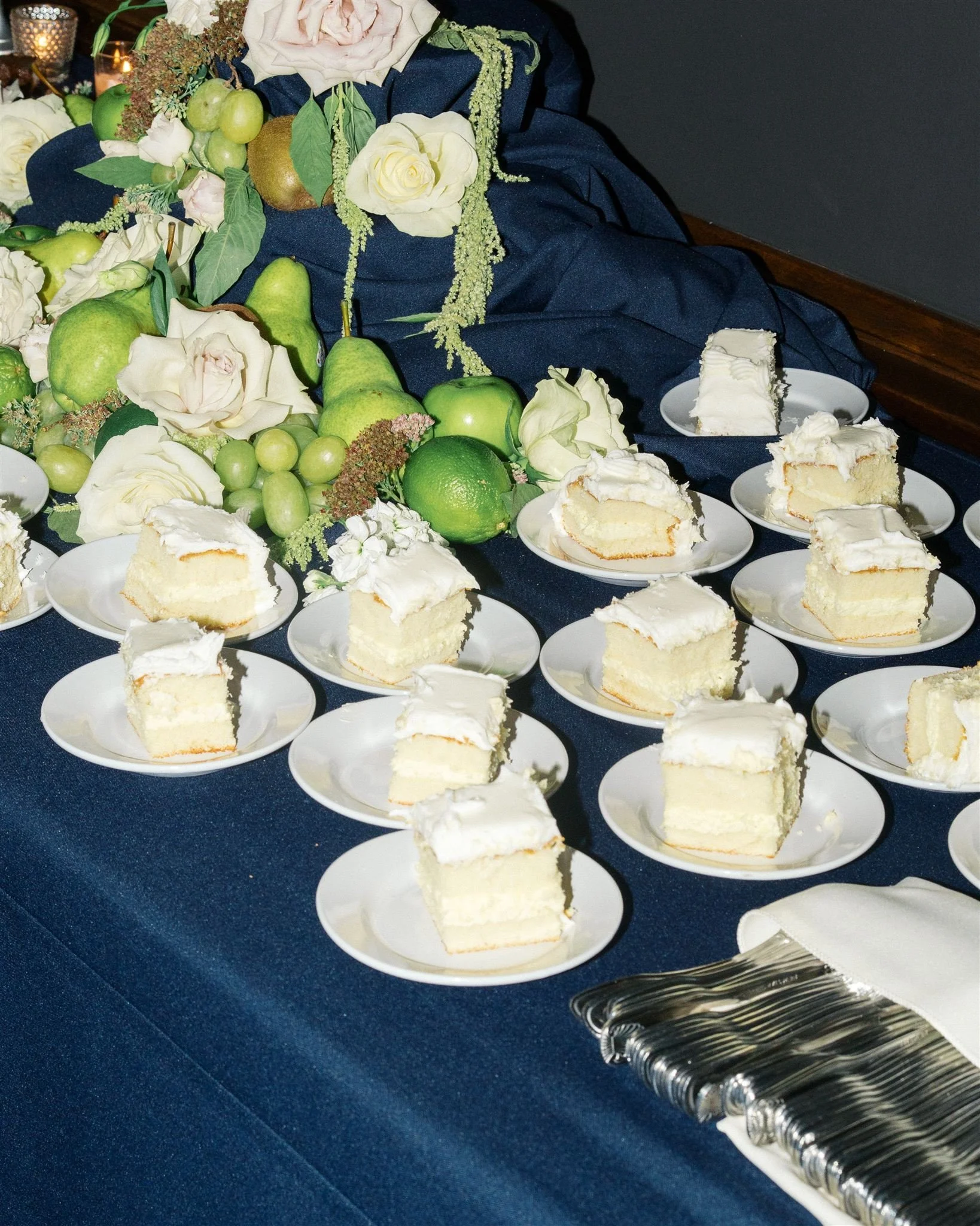 Slices of white frosted cake on small white plates arranged on a dark blue tablecloth, surrounded by green apples, pears, lime, white roses, and green leaves, with a bouquet of flowers and candles in the background at JPS Events at Regions Tower.  