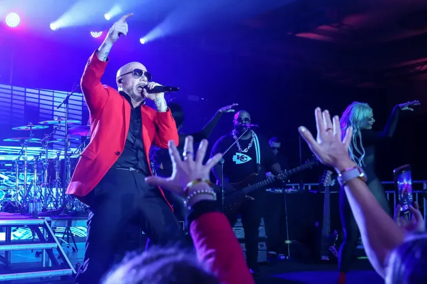 Performer wearing sunglasses and a red blazer singing into a microphone on stage with a band, audience reaching out, bright stage lights.