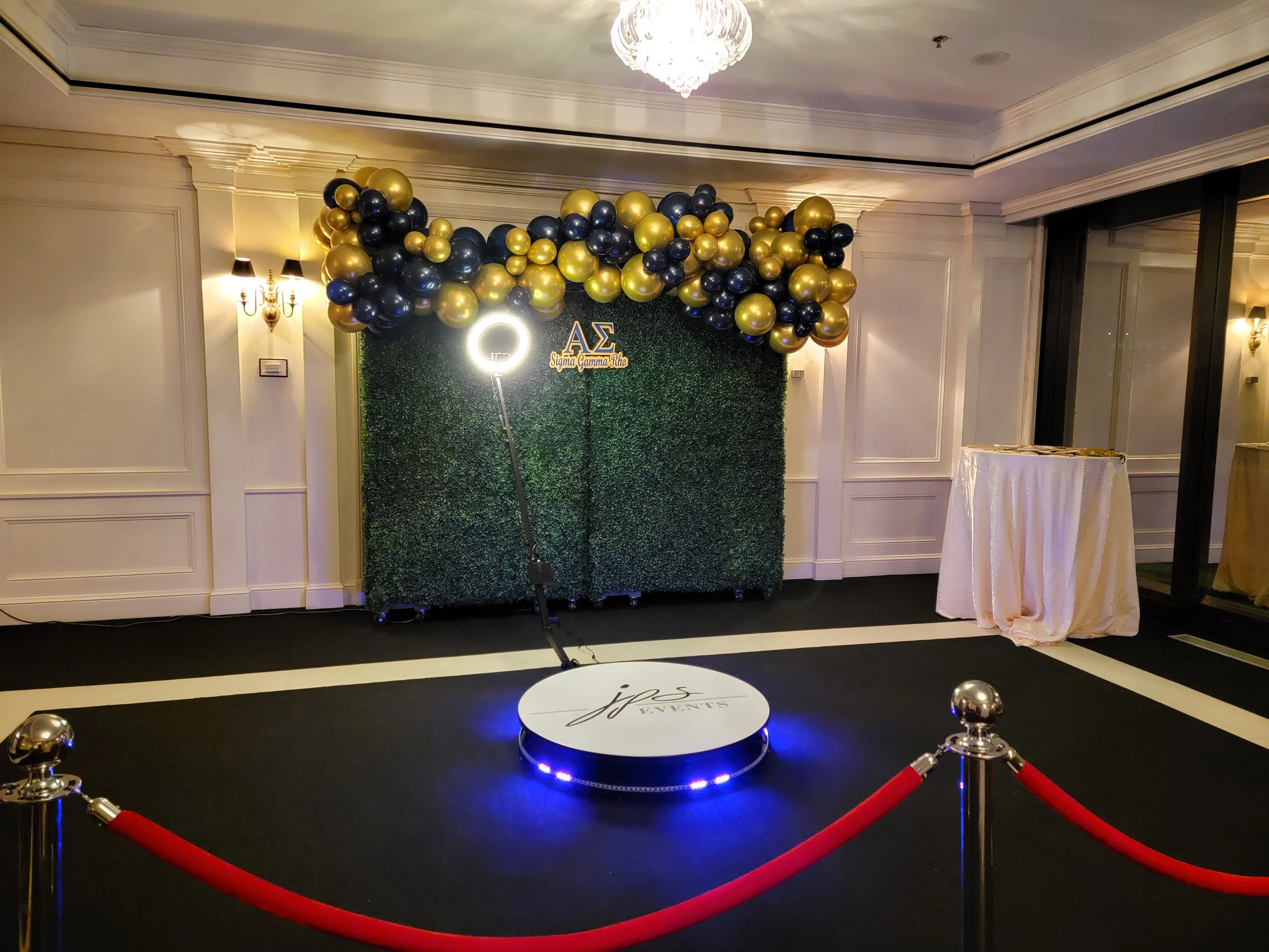 Sigma Gamma Rho corporate event photo backdrop at JPS Events at Regions Tower, featuring an in-house green foliage wall, 360 Photo Booth, custom illuminated neon branding, VIP red carpet ropes, and bespoke decor styling.