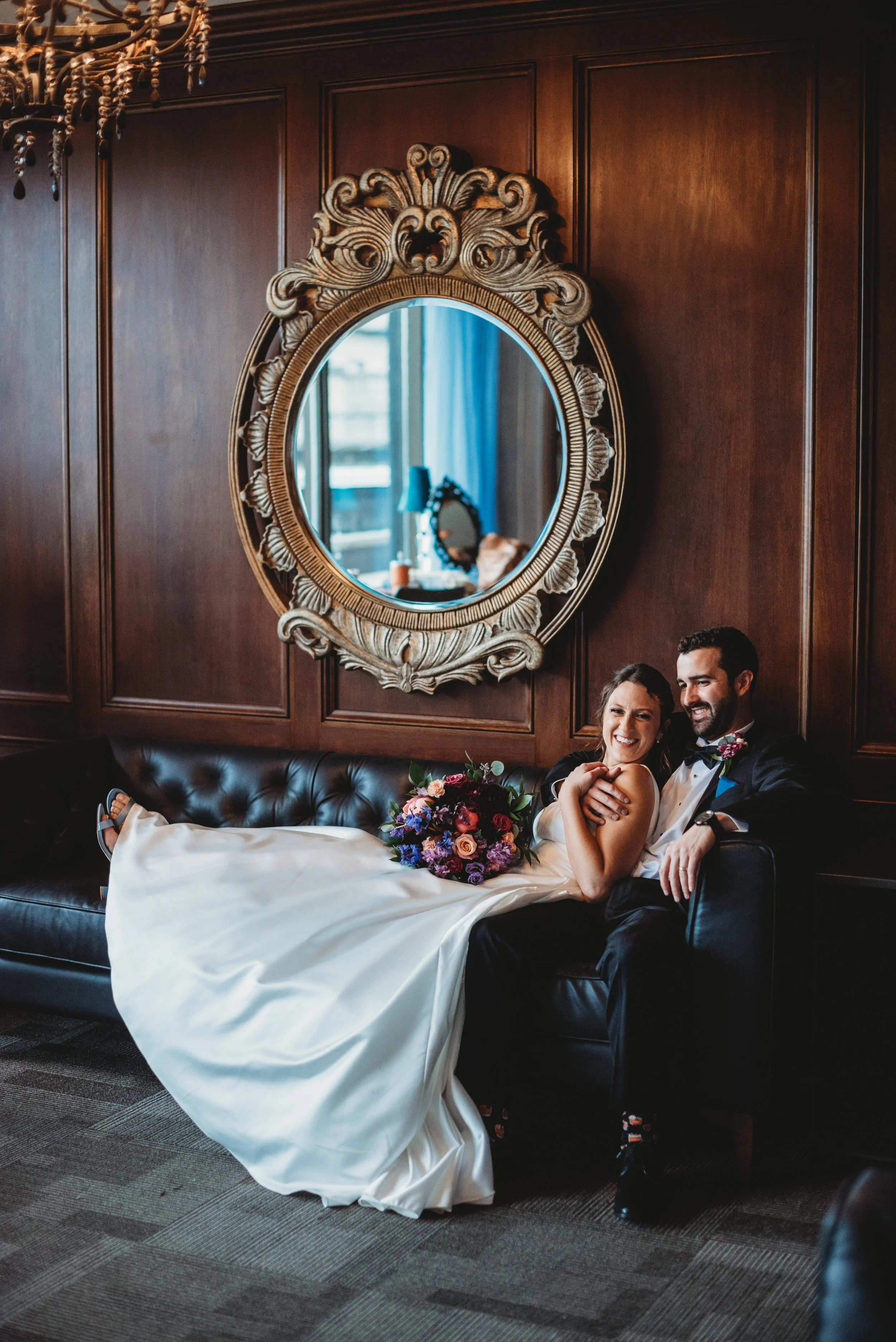 A bride and groom sitting on a black leather sofa in a wood-paneled room with an ornate gold mirror on the wall behind them. The bride is smiling, holding a bouquet of flowers, and leaning toward the groom, who is smiling and has his arm around her. 