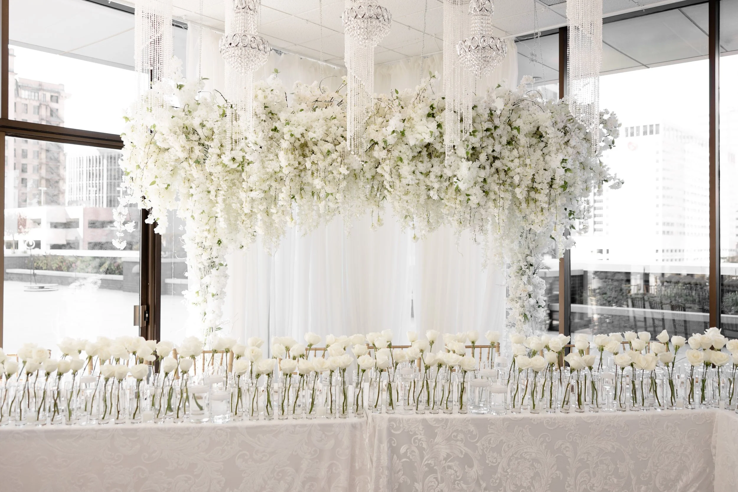 Custom white floral head table  arch designed by the in-house floral team at JPS Events at Regions Tower. Showcases our bespoke wedding design process, crystal chandeliers, and indoor floor-to-ceiling city views in downtown Indy.