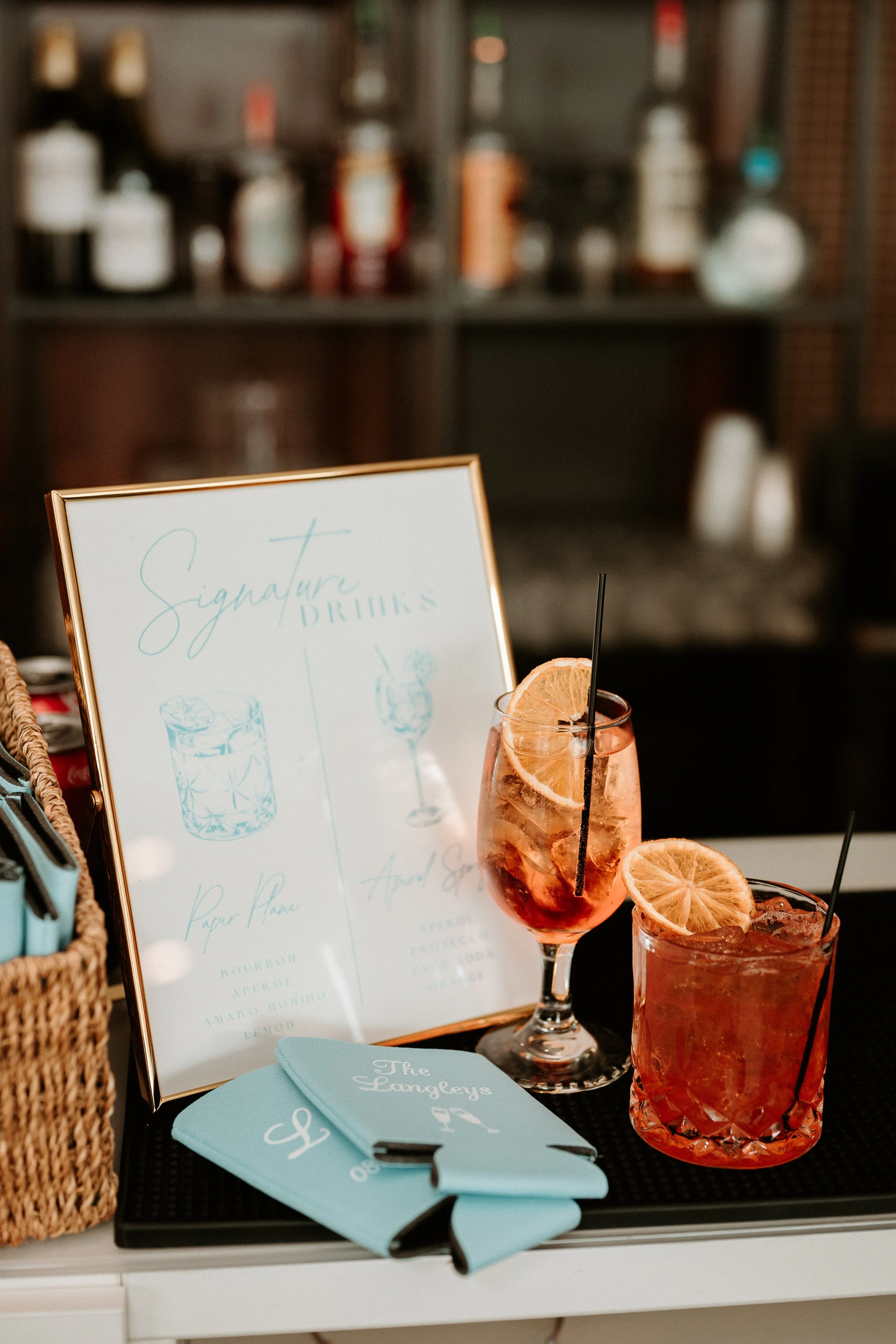 Two glasses of colorful cocktails with slices of citrus fruits on a bar counter, with a menu and a sign that reads 'Signature Drinks' in the background.