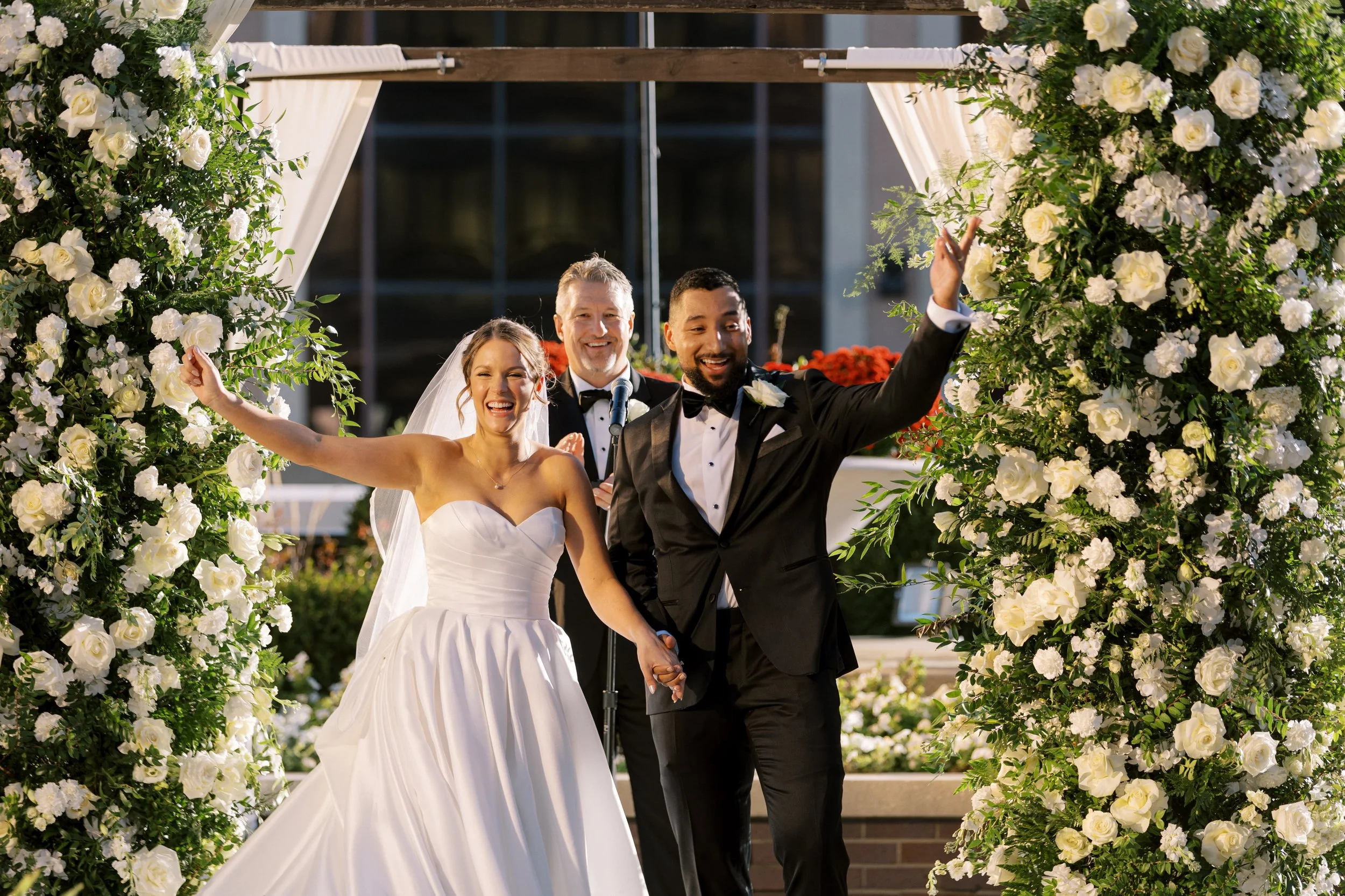A happy bride and groom celebrating their wedding as they walk hand in hand through a floral arch, with the groom raising his hand in celebration and all three women smiling joyfully.