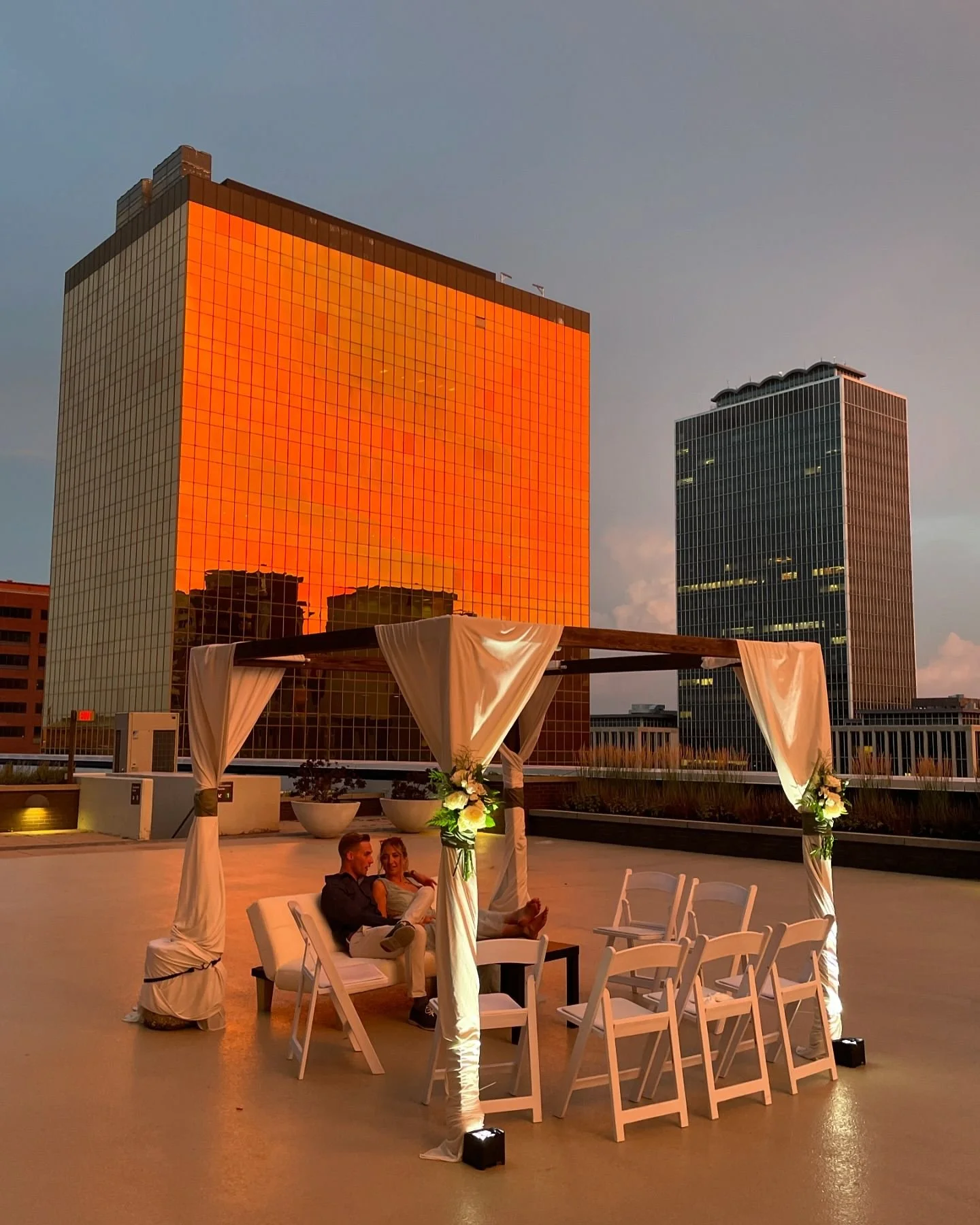 A rooftop wedding altar with white drapes and flowers, where a couple is seated, with city skyscrapers reflecting the orange sunset on the glass buildings in the background.