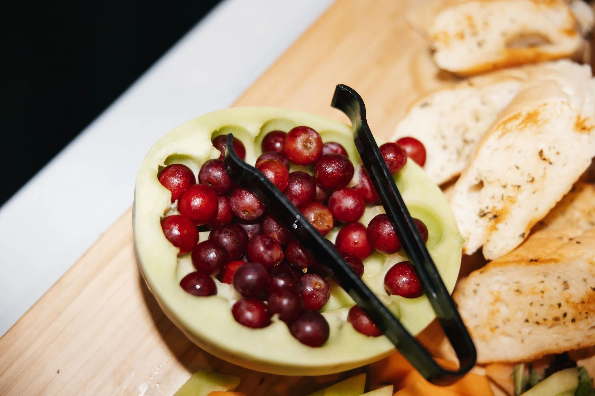 Red grapes inside a light green bell pepper with serving tongs, placed on a wooden cutting board next to sliced bread.