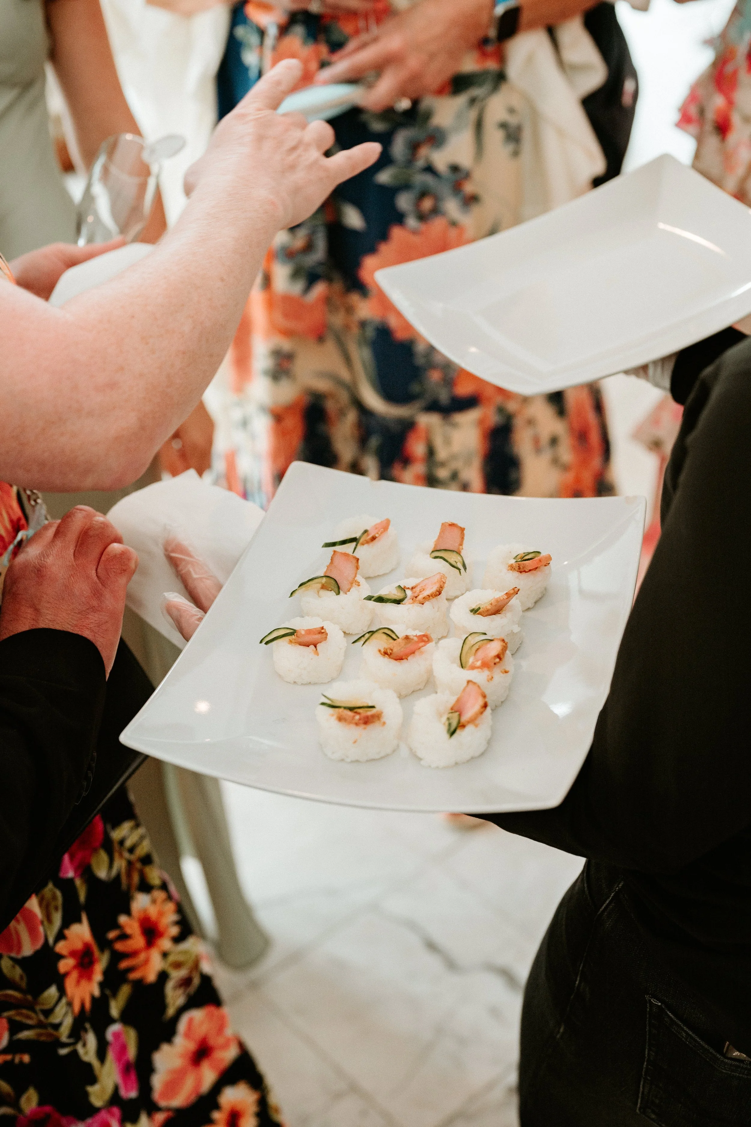 Close-up of a person holding a white square plate with sushi rolls topped with shrimp and cucumber. The person is wearing a black jacket. In the background, other people are visible, one with a floral dress and several with colorful clothing.