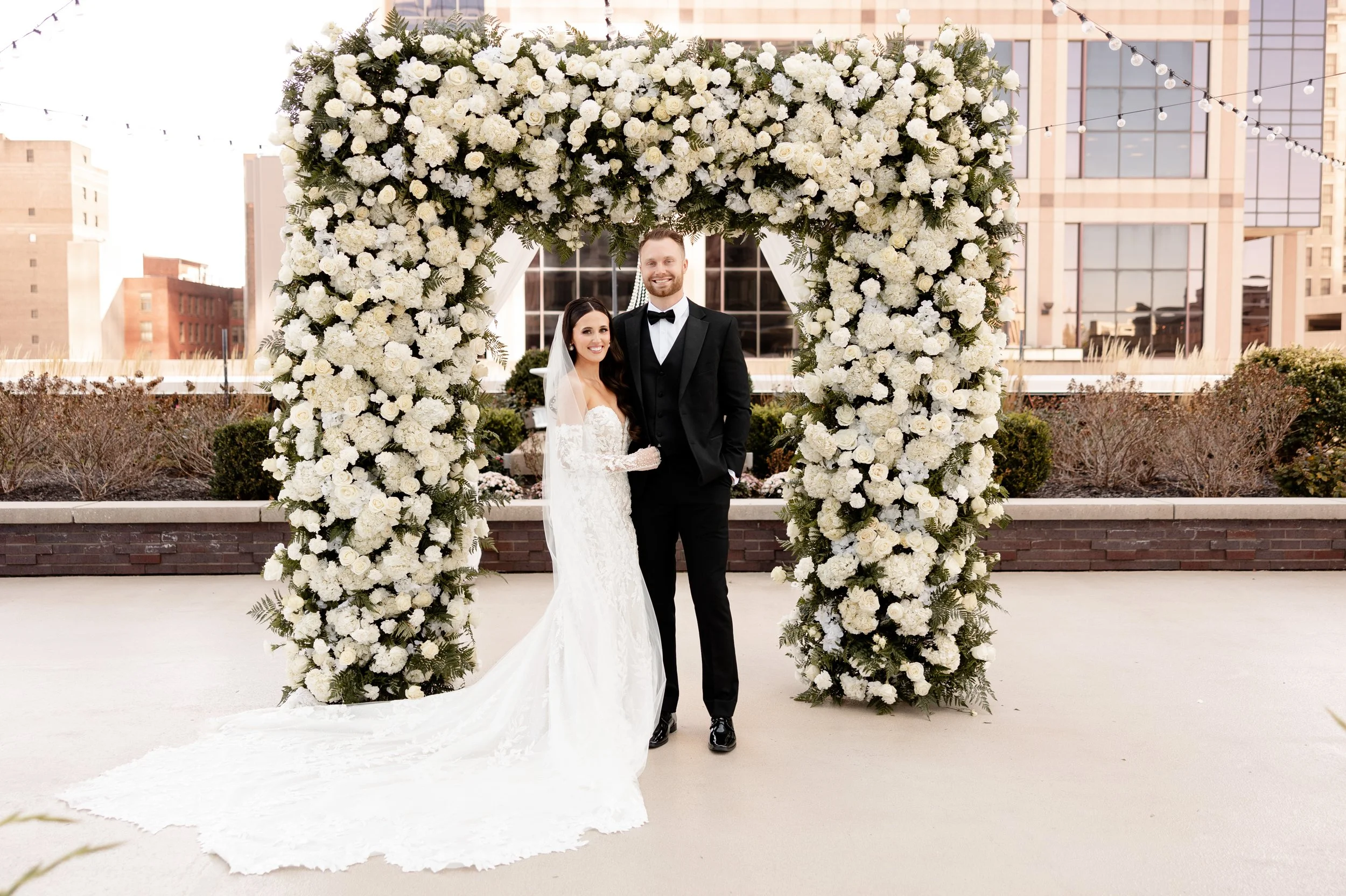Couple under a white rose floral arch on the 5th-floor rooftop terrace at JPS Events at Regions Tower. Designed by our in-house floral team, highlighting our all-inclusive custom design process and 360 Indy skyline views.