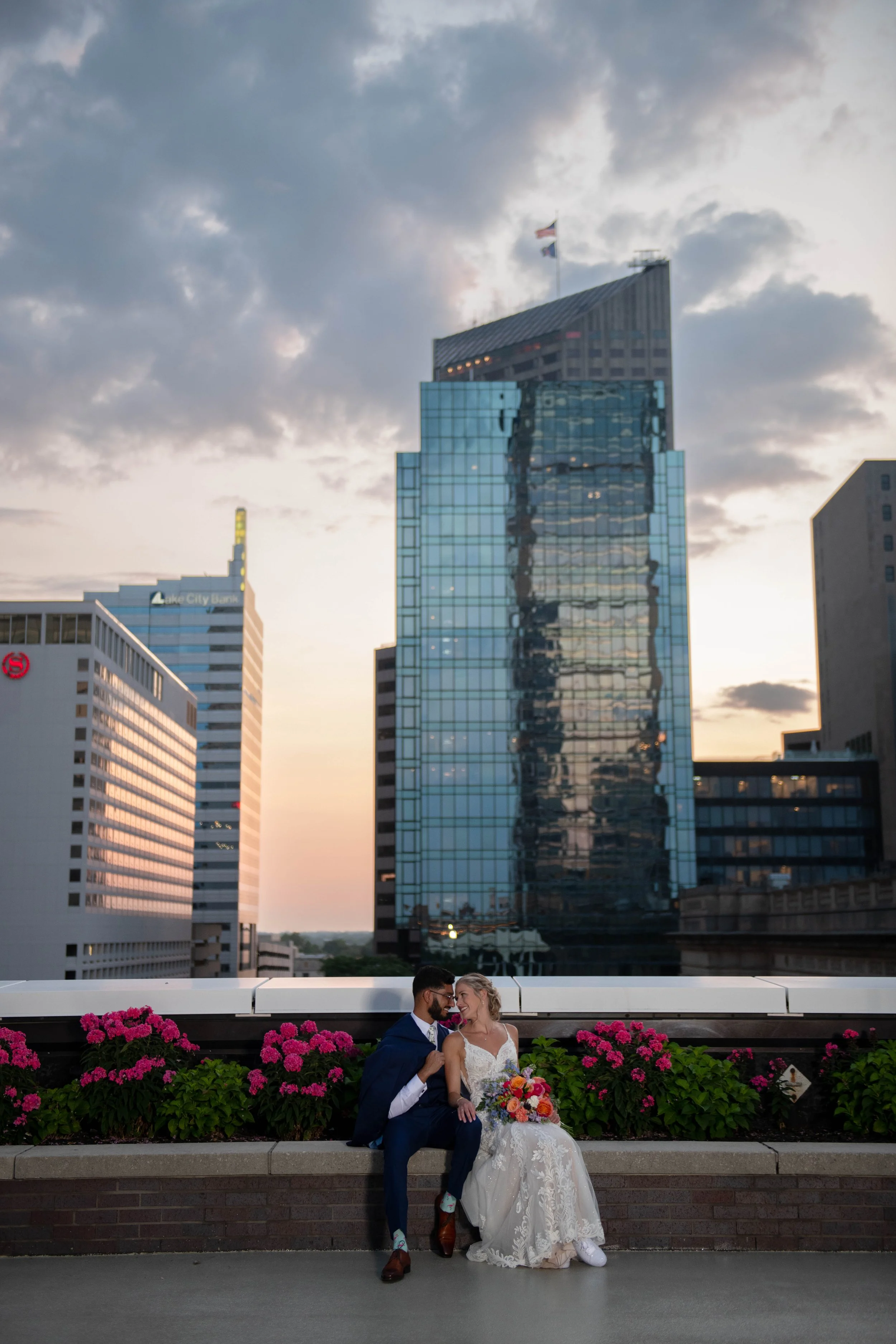 A couple in wedding attire sitting on a ledge with pink flowers in front of them, atop a building in an urban city skyline during sunset. The man is wearing a blue suit and the woman is wearing a white lace gown, holding a bouquet of colorful flowers