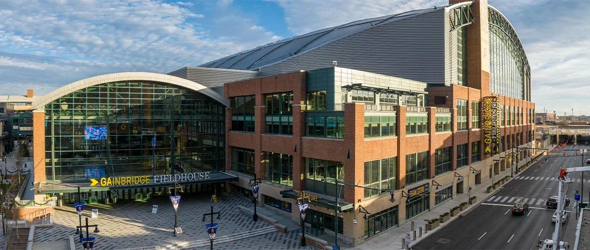 Exterior view of Gainbridge Fieldhouse, a large sports arena with glass and brick facade, multiple floors, and a curved roof, located beside city streets with cars and pedestrian crossings.