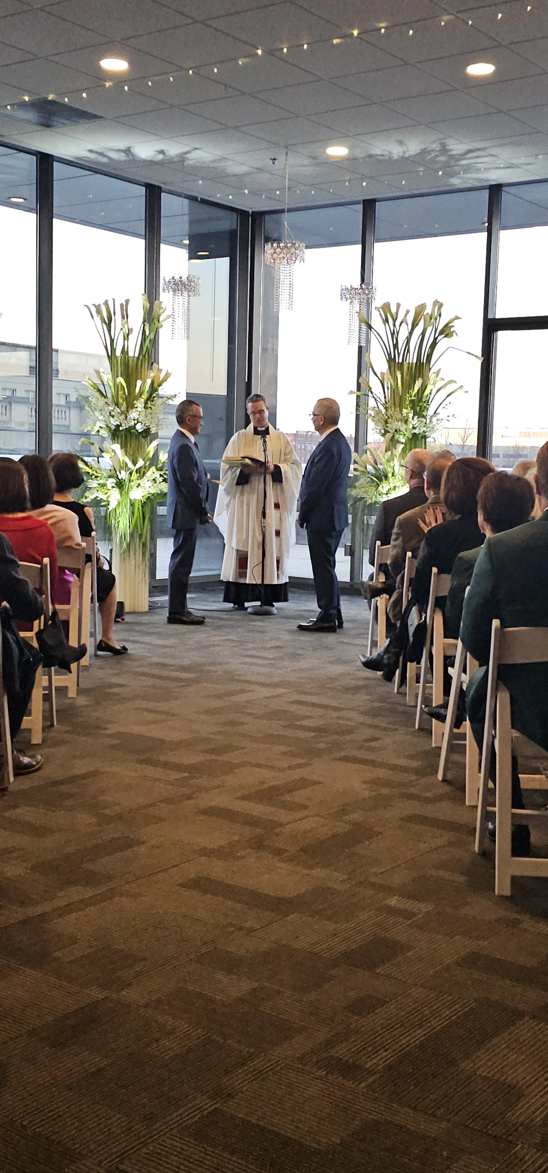 An lgbt friendly indoor wedding ceremony inside the modern luxury ballroom at JPS Events at Regions Tower in Downtown Indianapolis, featuring floor-to-ceiling skyline windows, chandeliers, and custom in-house floral design.