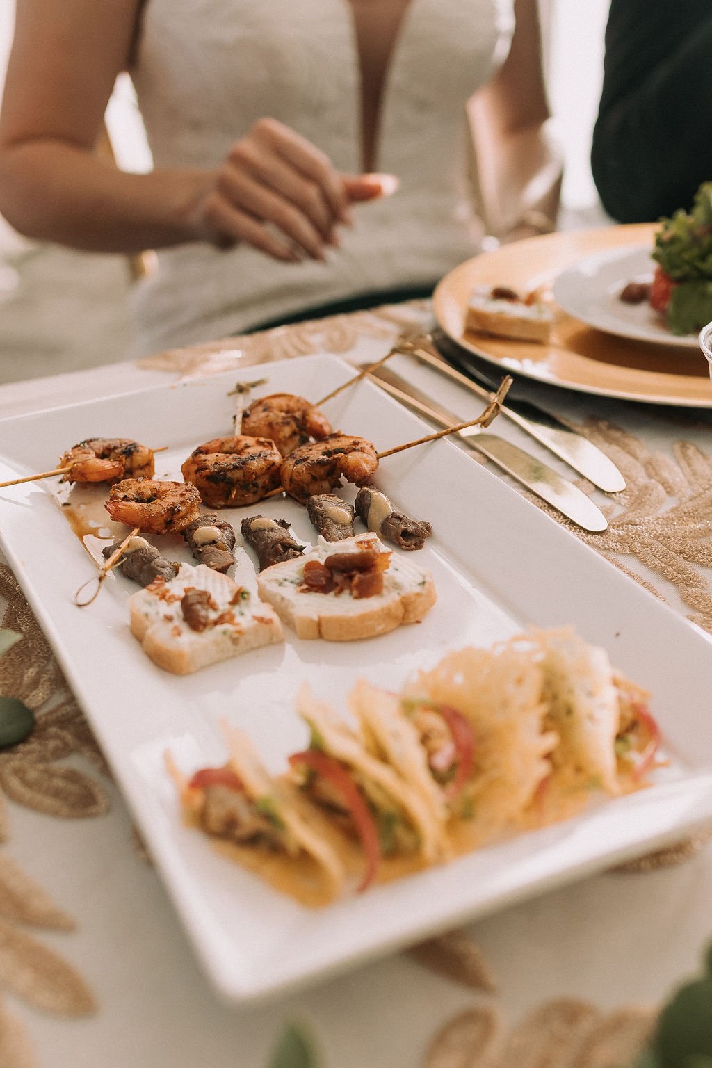 Close-up of a white rectangular plate with shrimp skewers, canapés topped with meat and mushrooms, and small sandwiches, on a table with cutlery and other plates in the background.