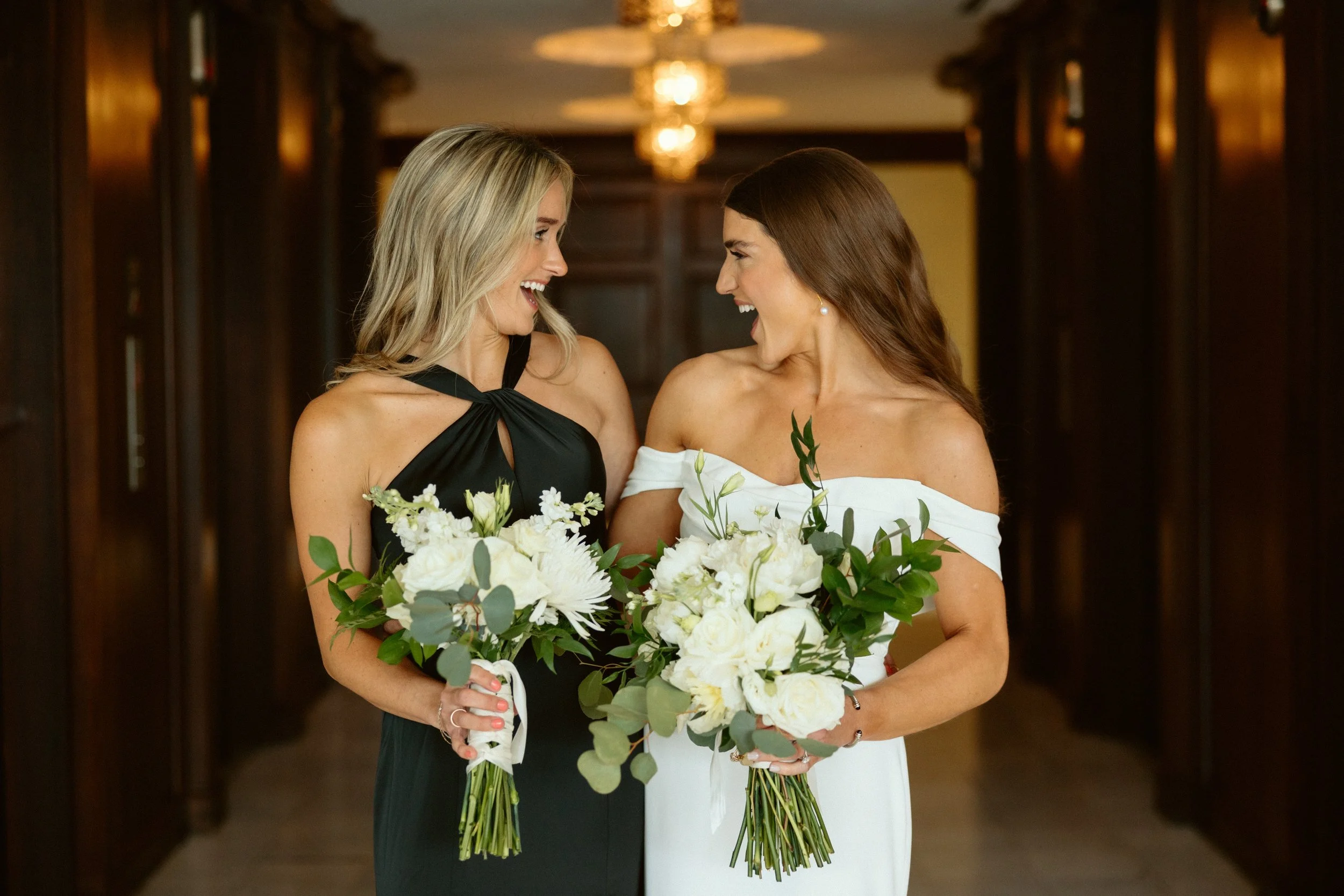 Bride and bridesmaid with custom white bouquets in the private suite hallway at JPS Events at Regions Tower. Highlights dark wood paneling, in-house wholesale floral design, & family-owned coordination in downtown Indy.