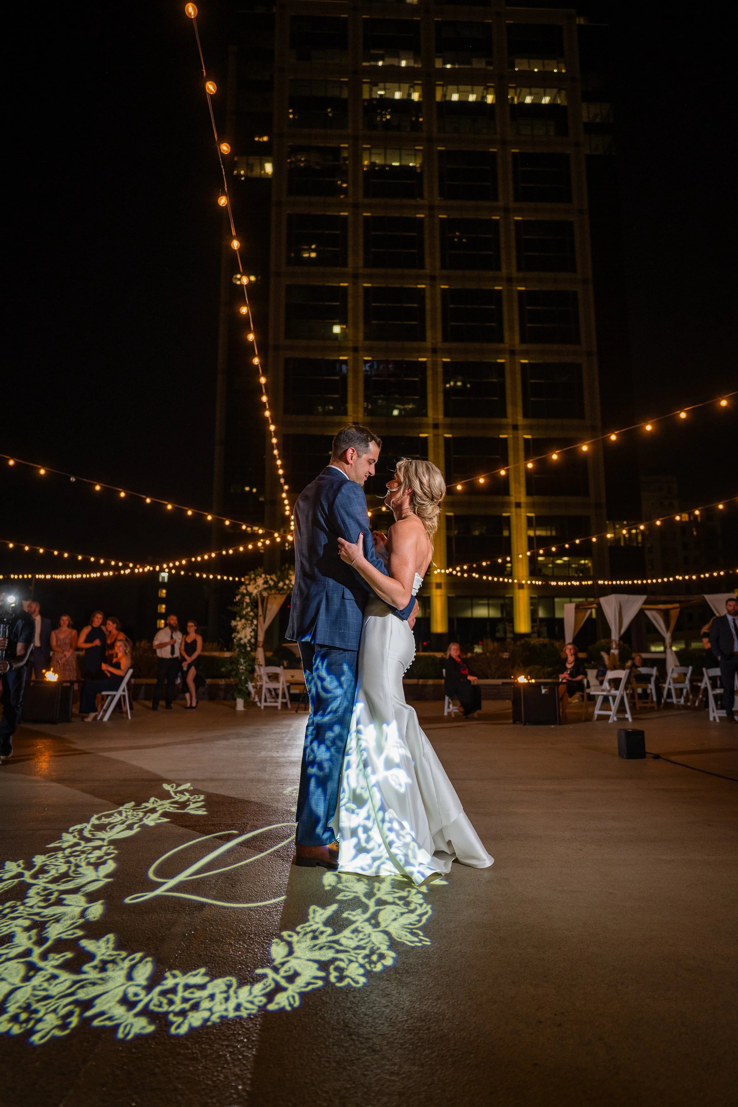 A bride and groom are dancing on an outdoor wedding dance floor at night, illuminated by string lights and a projected floral design on the ground. The bride is wearing a strapless white gown, and the groom is in a blue plaid suit. Guests are seated 