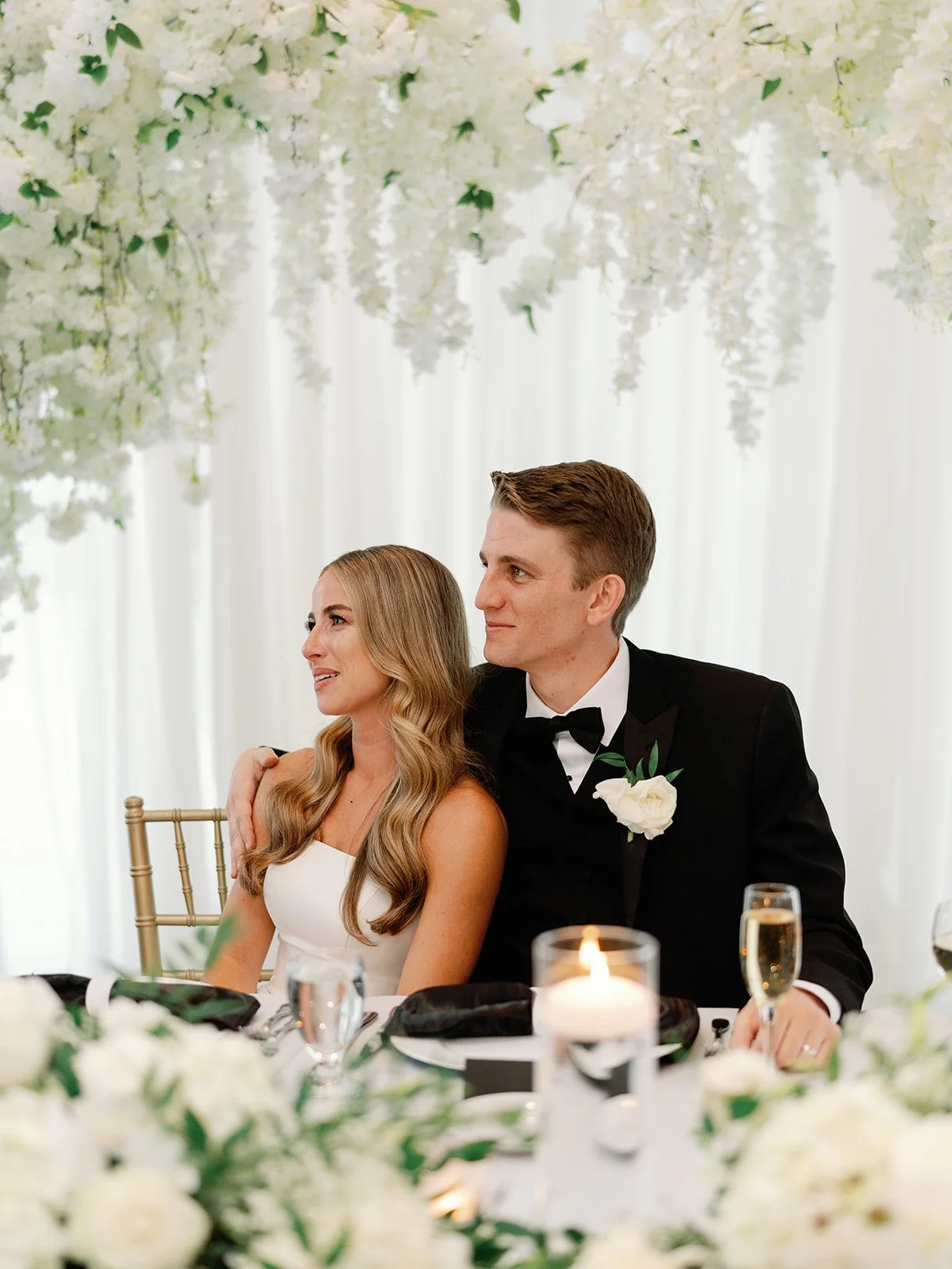 A bride and groom sitting together at a wedding reception table decorated with white flowers and candles. The bride has long wavy blonde hair and is wearing a white dress, while the groom has short brown hair and is dressed in a black tuxedo with a w