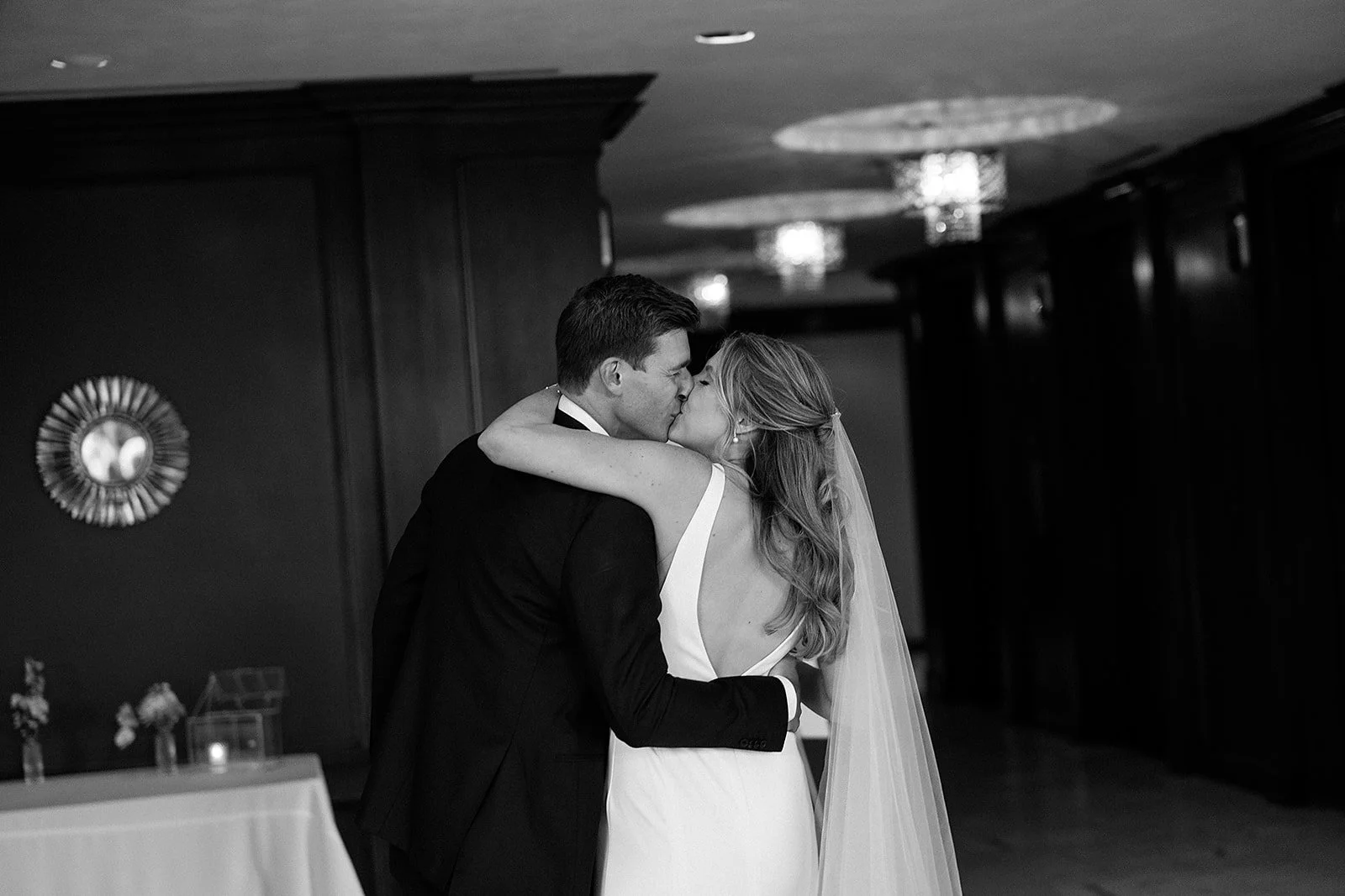 A black and white photo of a bride and groom sharing a kiss during their wedding reception inside a decorated venue with hanging lights.