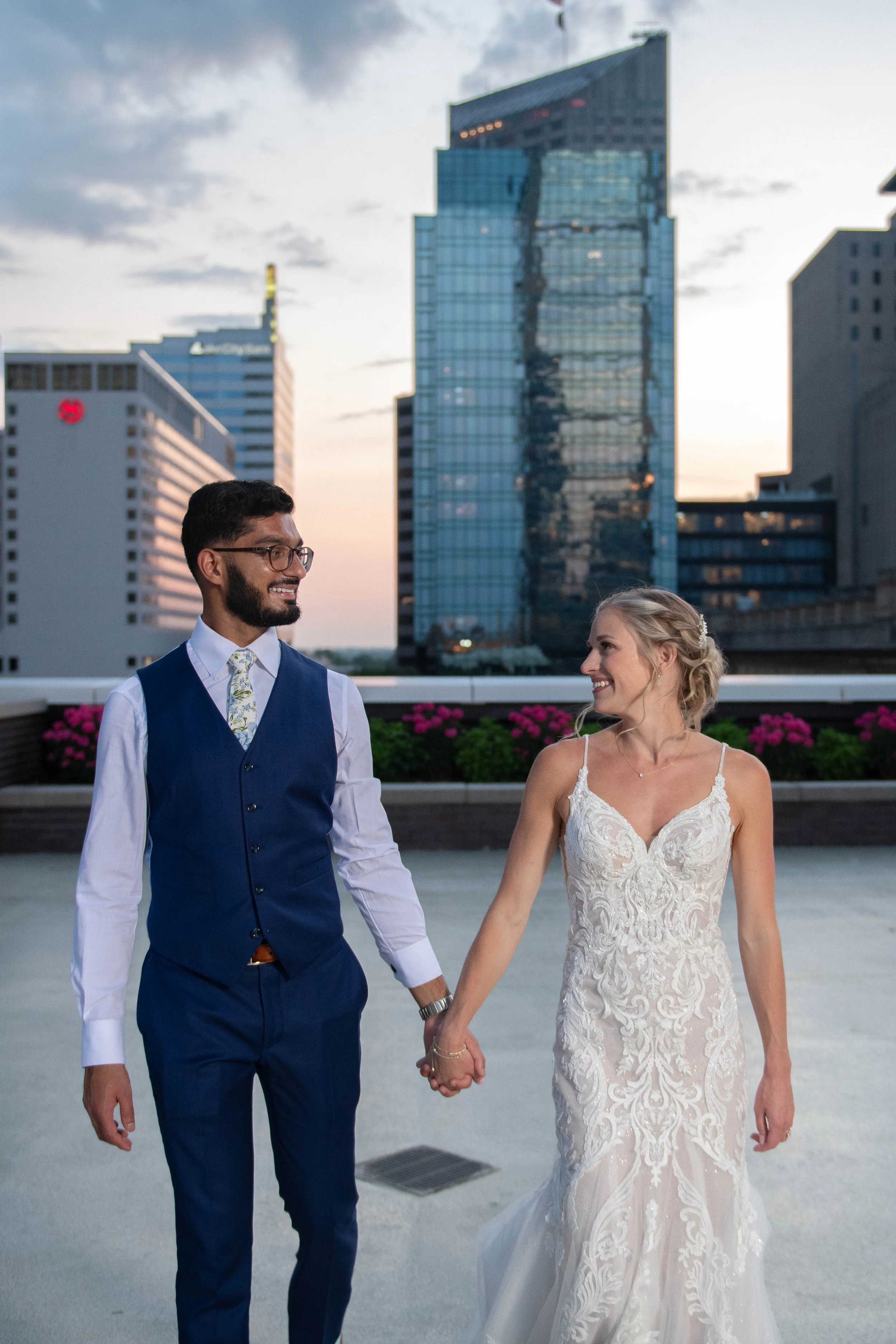 A newlywed couple holding hands and smiling at each other on the JPS Events' private/exclusive rooftop in downtown Indy during dusk, with city skyscrapers in the background.