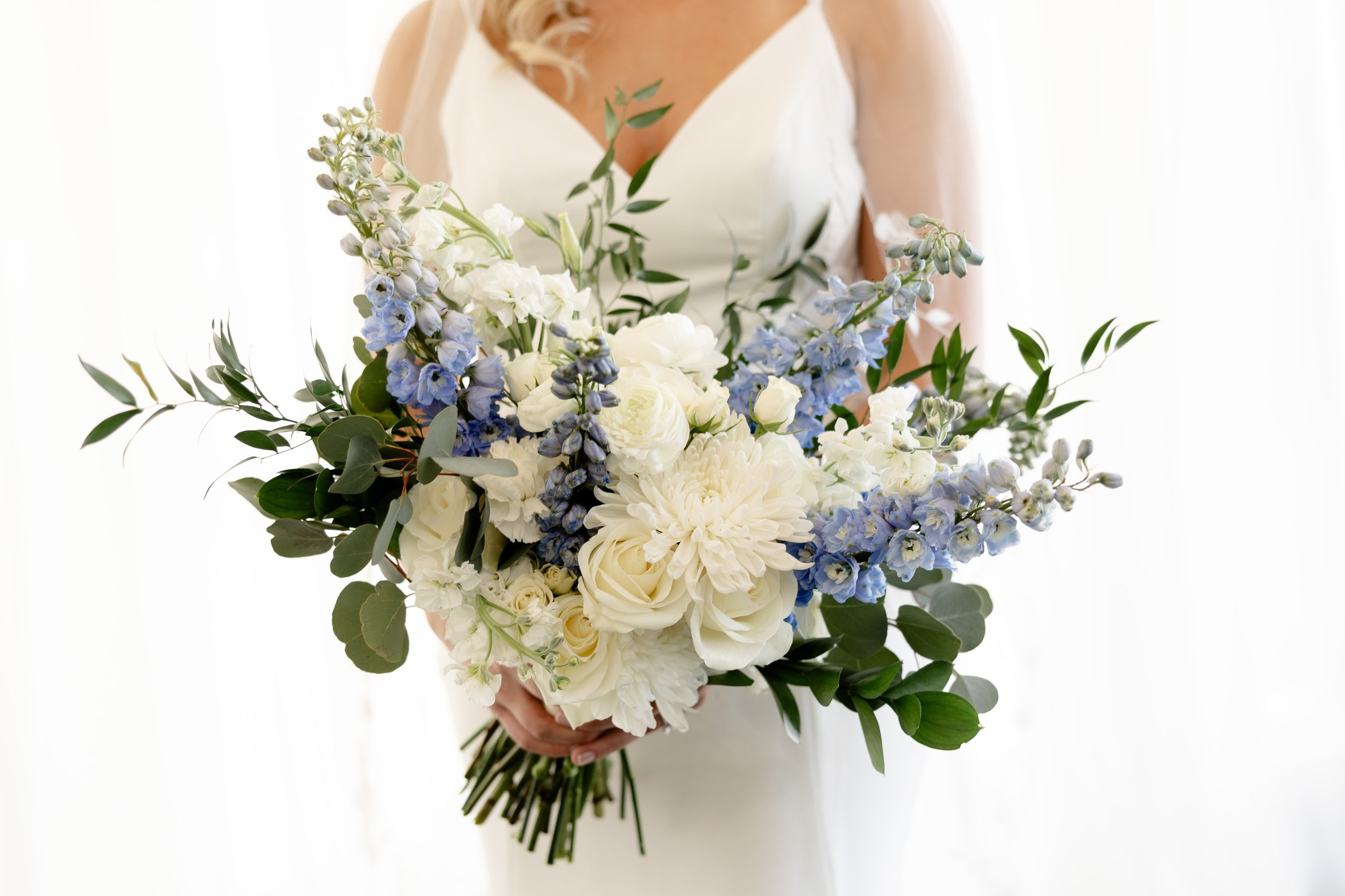 A woman in a white dress holding a large bouquet of white and blue flowers with greenery against a white background.