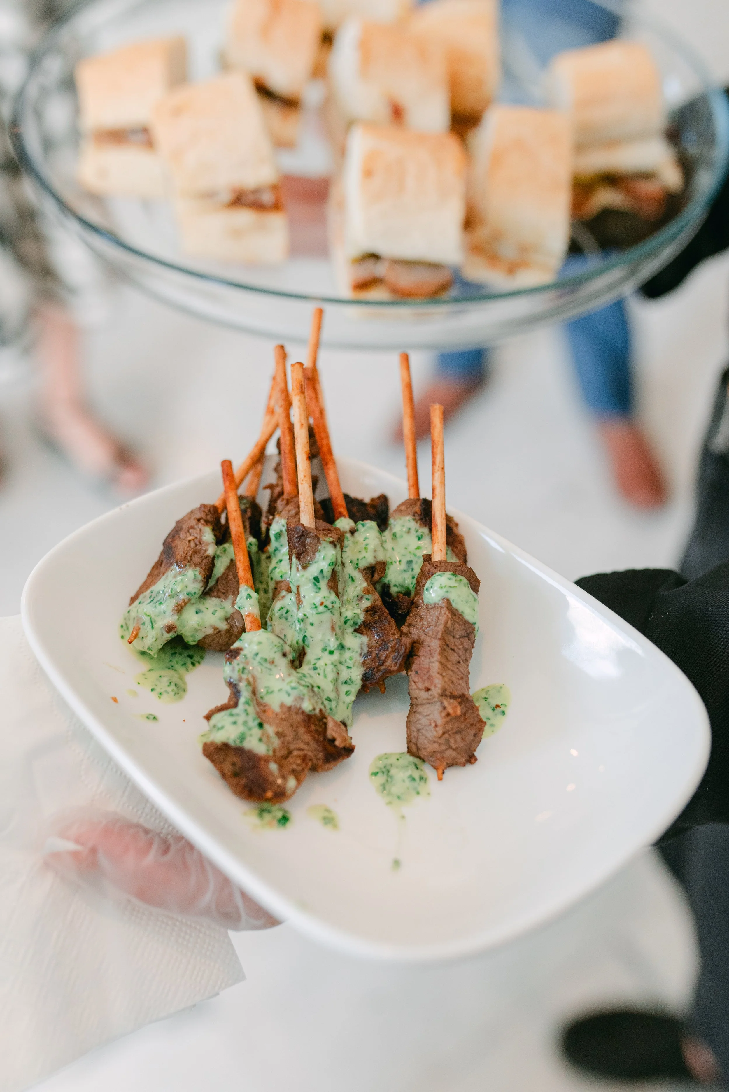 Close-up of a white bowl containing grilled meat skewers topped with green herb sauce, with a blurry glass cake stand holding sandwiches in the background—passed hors d'oeuvres by luxury in-house catering at JPS Events at Regions Tower in Indy.