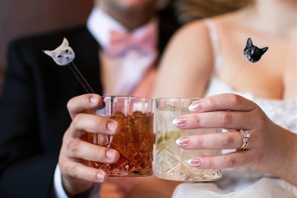 A couple holding cocktails, with animal-shaped drink stirrers with cat faces, celebrating an event. The woman wears a wedding ring.