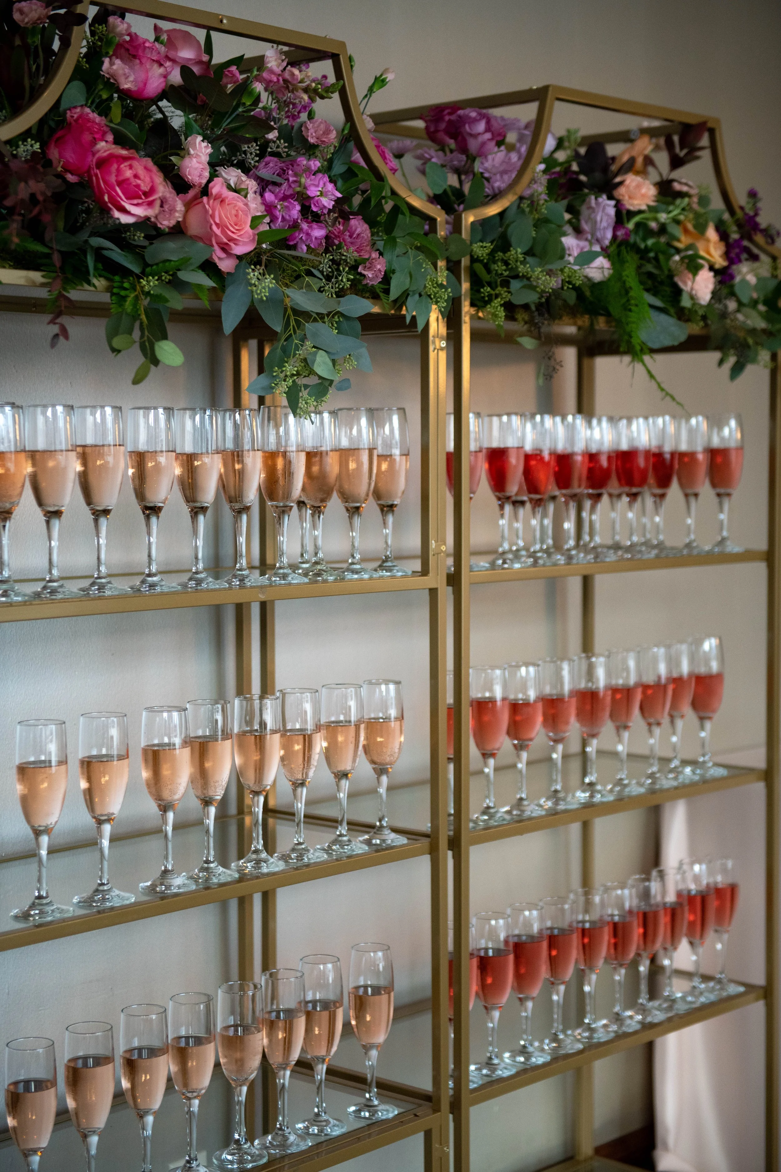 A gold-colored shelving unit with multiple shelves holding rows of champagne glasses filled with pink and red drinks. The top shelf is decorated with a lush arrangement of pink and purple flowers and greenery.