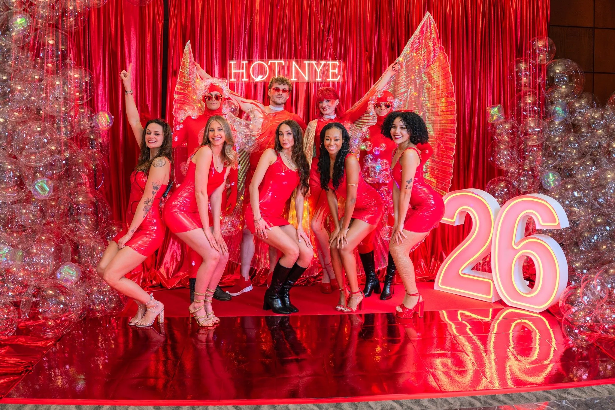 Group of people in red outfits and accessories, standing in front of a red curtain with balloons and a neon sign that says 'HAPPY NEW YEAR 2023'.