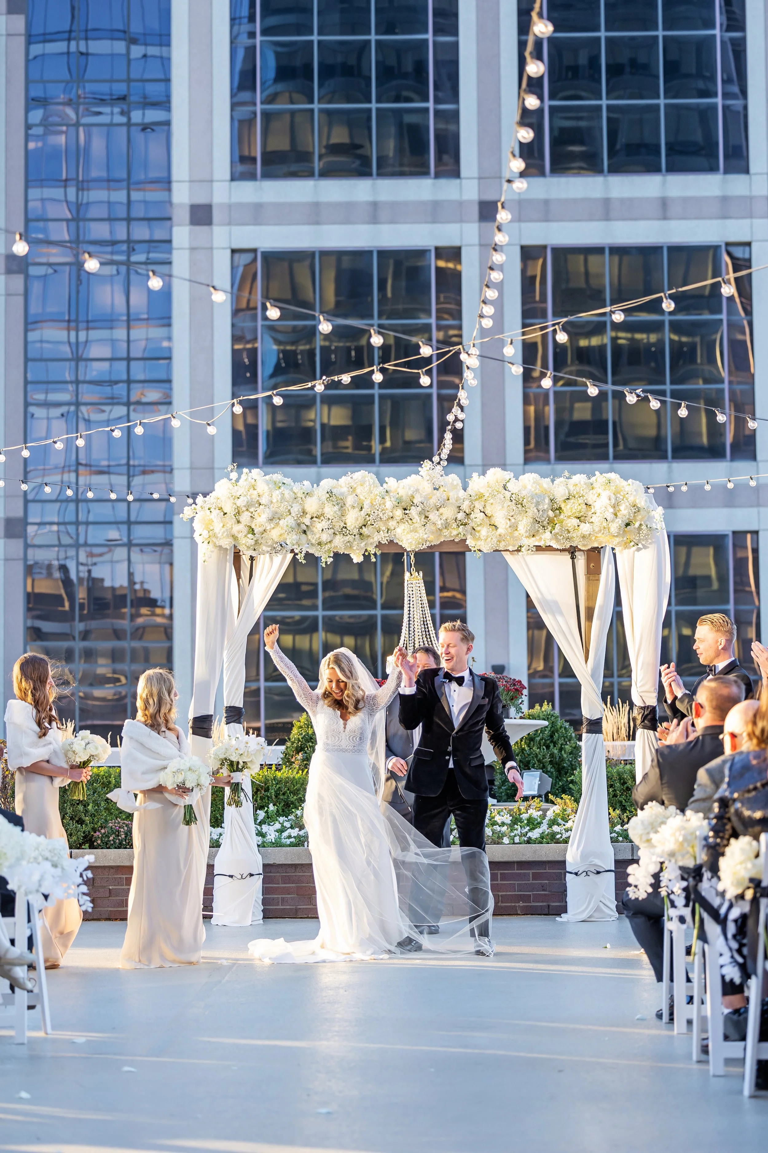 A wedding ceremony outdoors with a bride and groom, surrounded by guests, under a decorated canopy with white flowers and string lights, in front of a modern glass building.