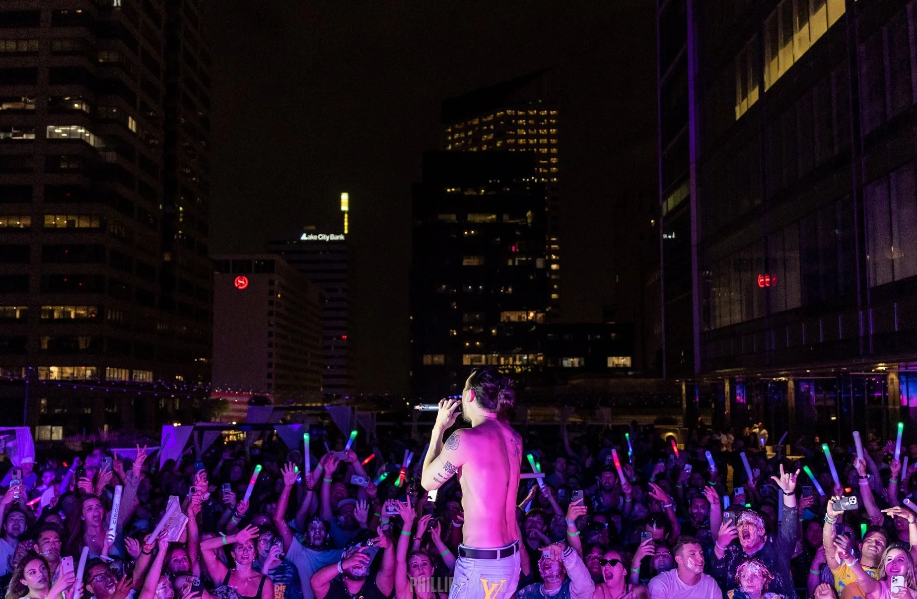 A shirtless performer with purple hair and tattoos on their arm on a stage in front of a large crowd at night in an urban setting, with tall buildings and city lights in the background. The crowd is excited, holding glowing sticks and raising their hands.