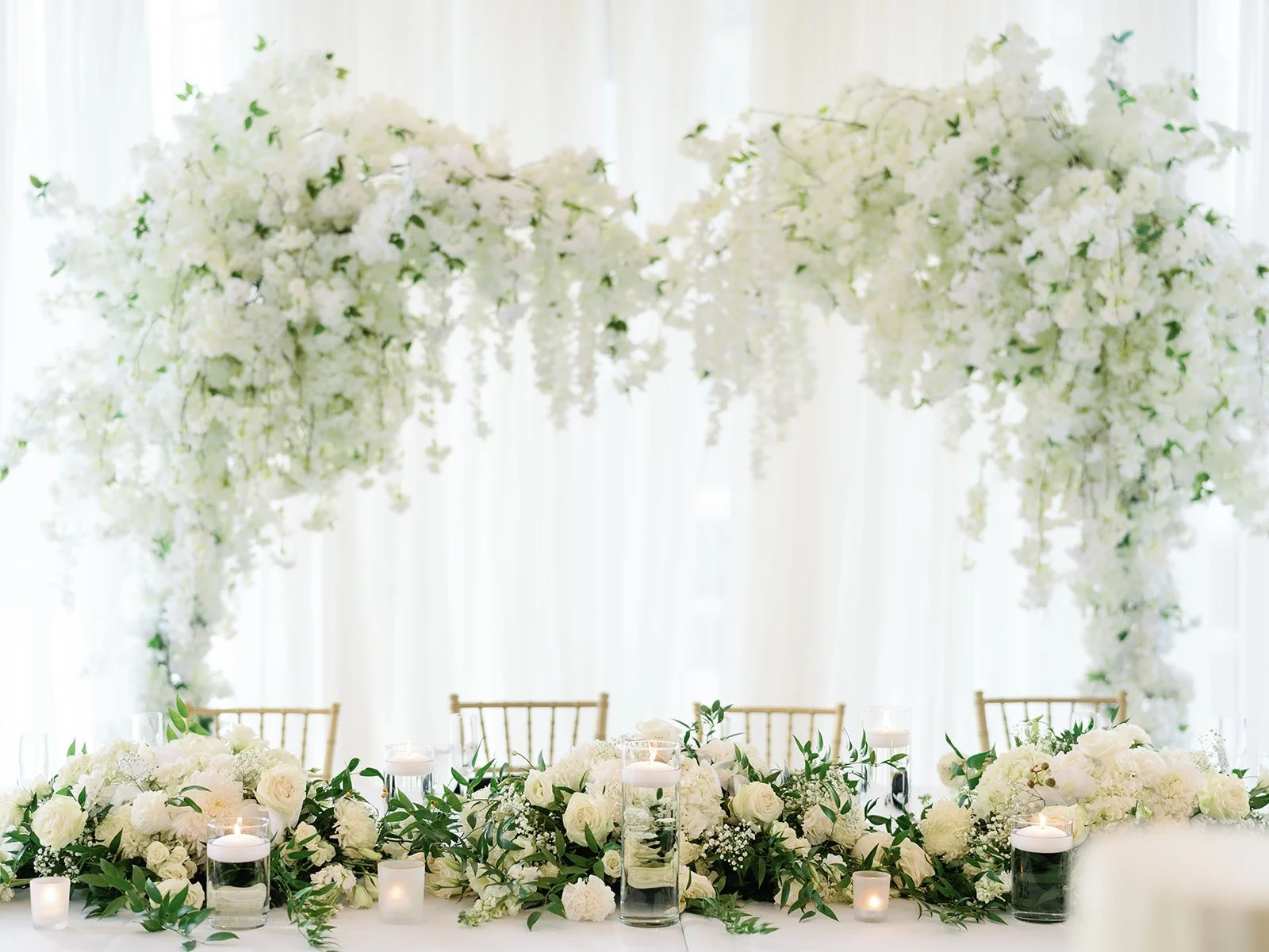 Elegant wedding table decorated with white roses, greenery, candles in glass holders, and a floral arch overhead with white flowers and greenery in the background.