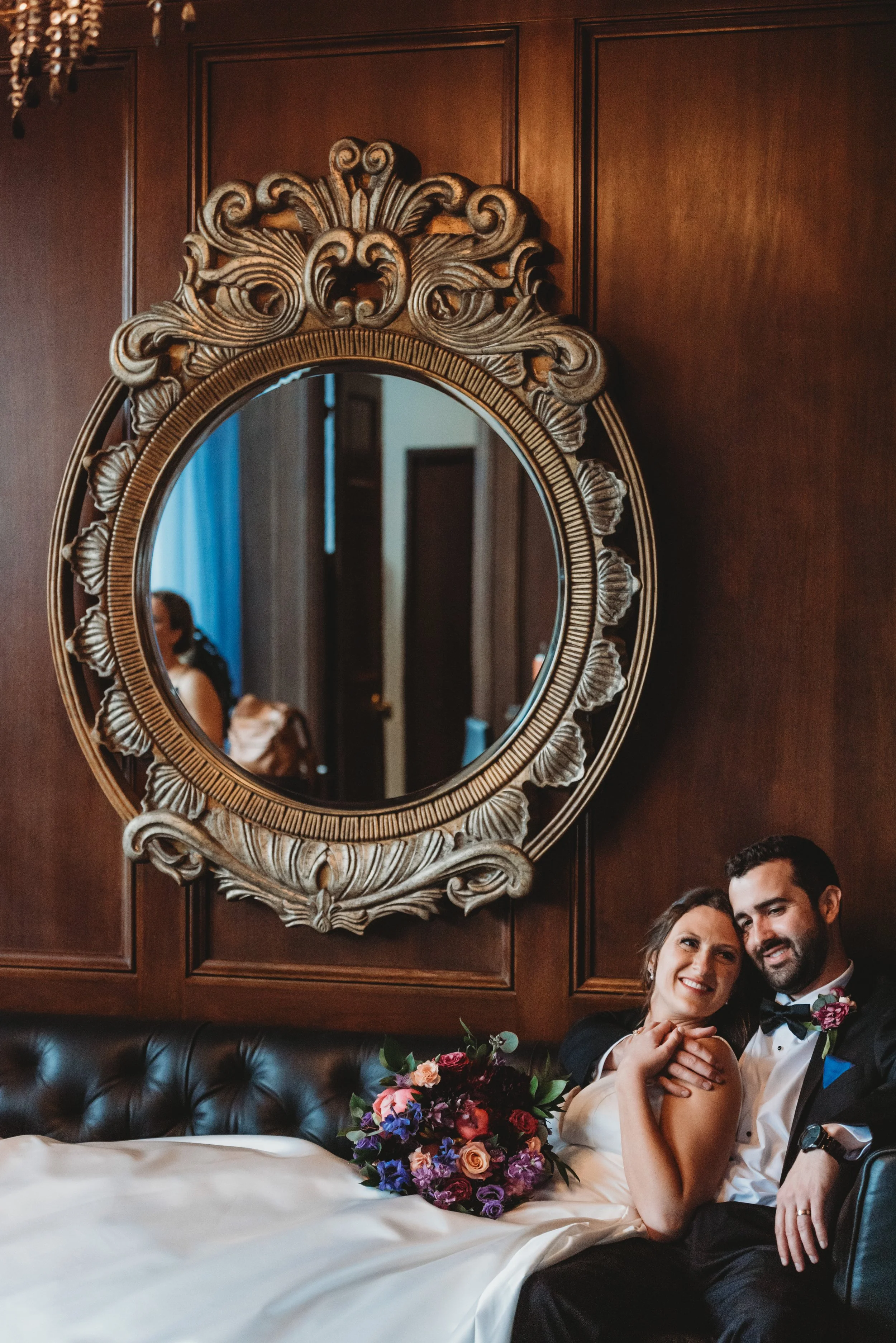 A couple dressed in wedding attire, sitting on a leather bench in a room with dark wood paneling, a large ornate mirror behind them, and a bouquet of colorful flowers placed in front of them.